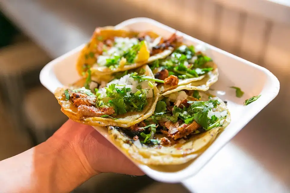 A hand holding a foam tray of al pastor tacos topped with chopped onion and coriander