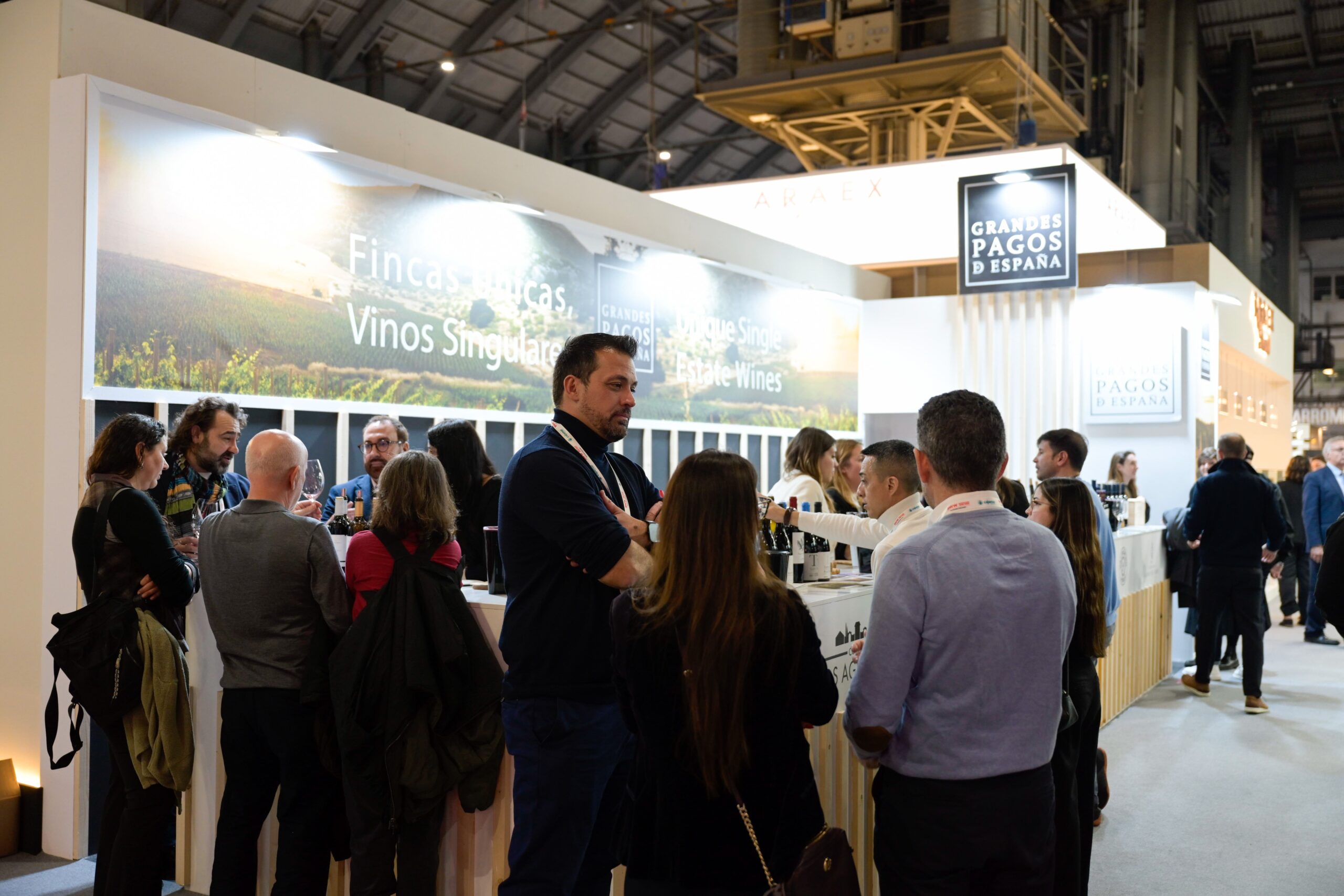 Visitors tasting wine at branded exhibitor booths on the Barcelona Wine Week exhibition floor, with 'Grandes Pagos de España' and 'Fincas Únicas, Vinos Singulares' signage overhead