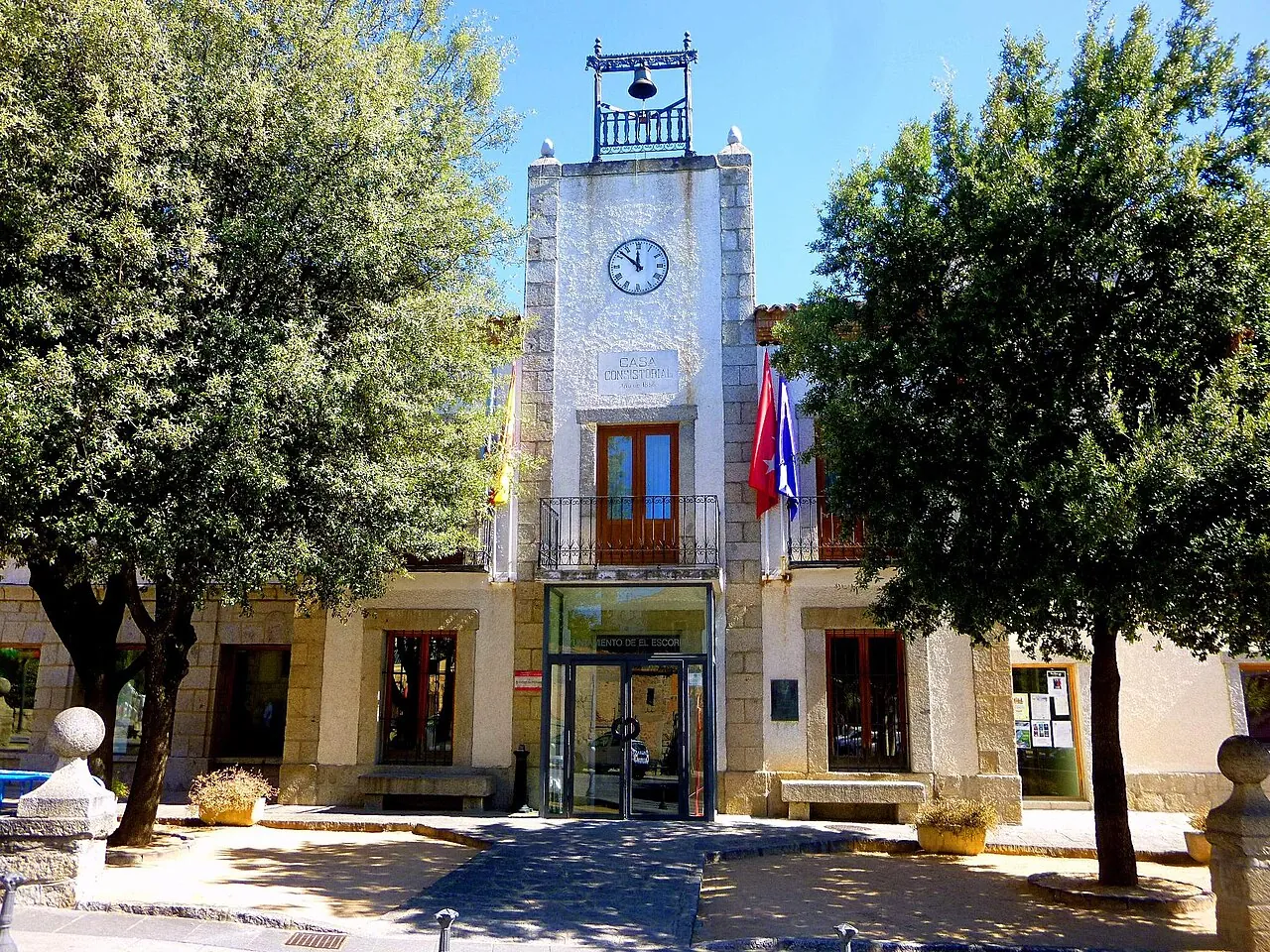 The town hall of El Escorial, a stone and white building with a clock and bell tower, framed by trees