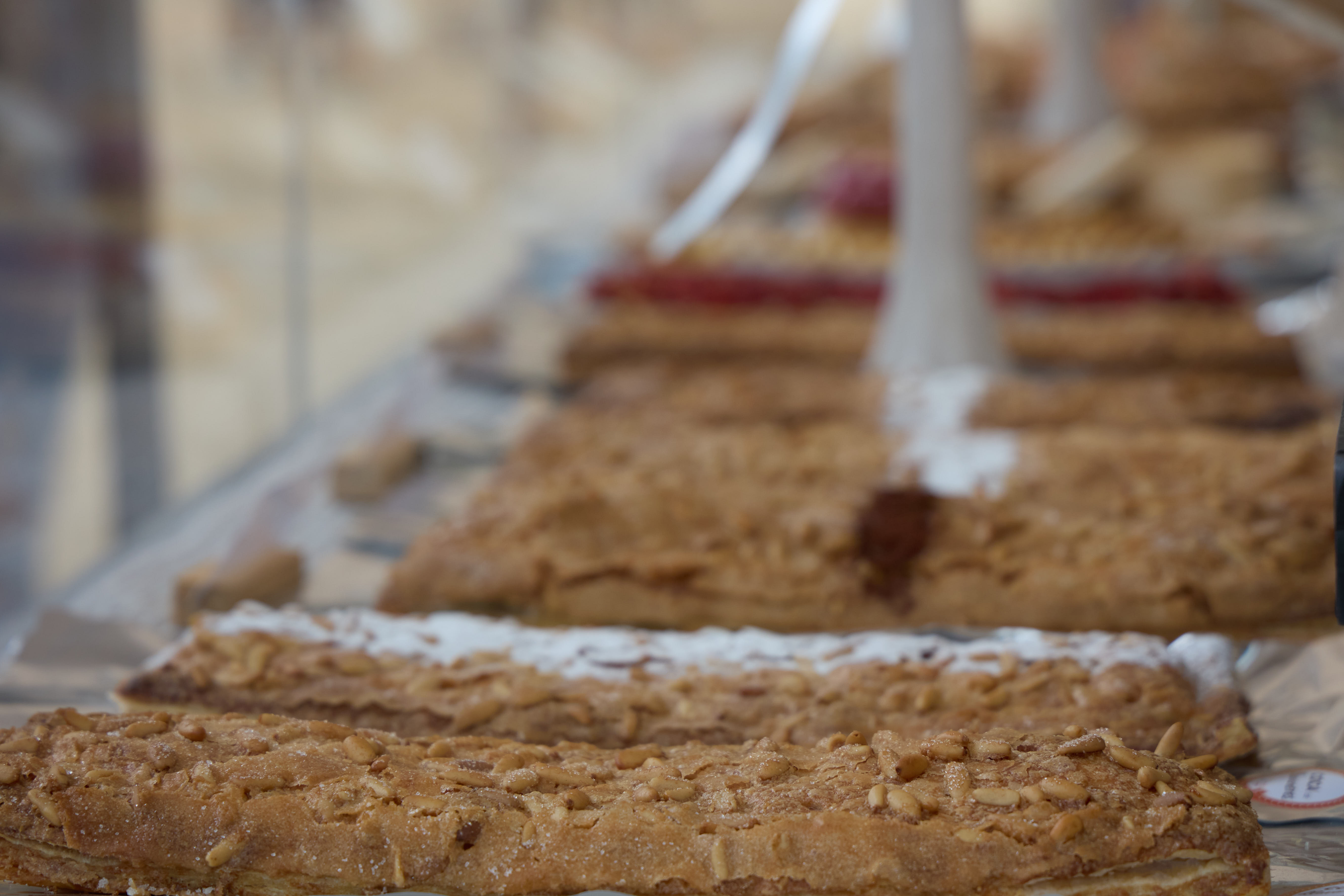 Close-up of rectangular cocas topped with pine nuts and sugar on a market table, more pastries blurred behind (2025 edition)