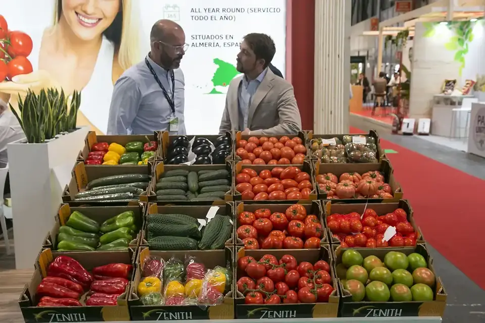 Crates of tomatoes, peppers, cucumbers and courgettes on display at an exhibitor stand, with two visitors talking behind