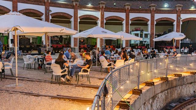 Visitors seated under white umbrellas at dusk in the courtyard of the Museu del Ferrocarril de Catalunya during Gastrofest, with vintage locomotives visible in the roundhouse behind them