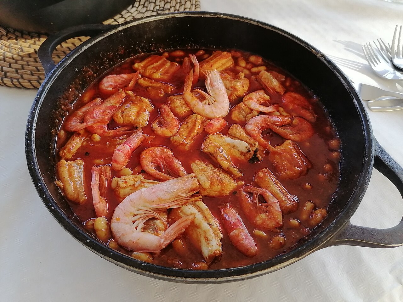 A black cast-iron pan of Tarragona-style romesco fish stew with prawns, fish pieces and white beans in a red sauce
