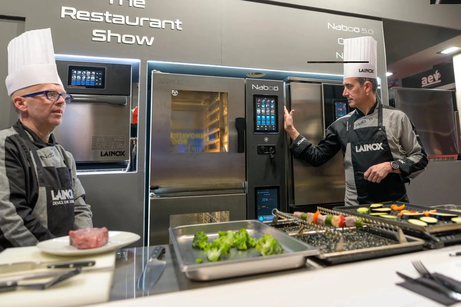 Two chefs in whites demonstrating a combination oven at a stand, with grilled vegetables and meat on trays
