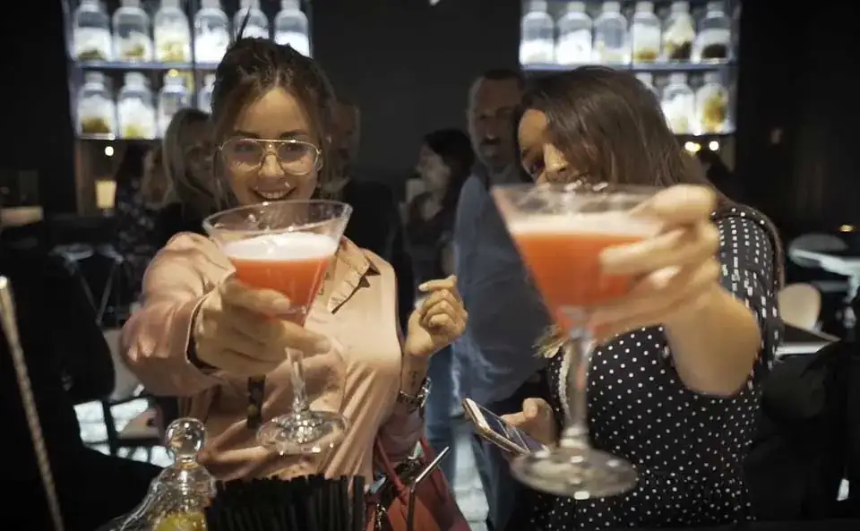 Two women holding up pink cocktails in coupe glasses at a bar