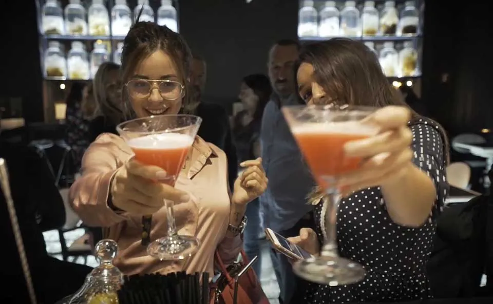 Two women holding up pink cocktails in coupe glasses at a bar