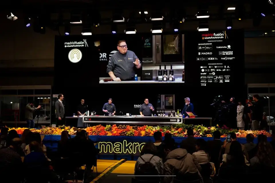 A chef cooking on the Madrid Fusión auditorium stage in front of a seated audience, with crates of vegetables along the front of the stage
