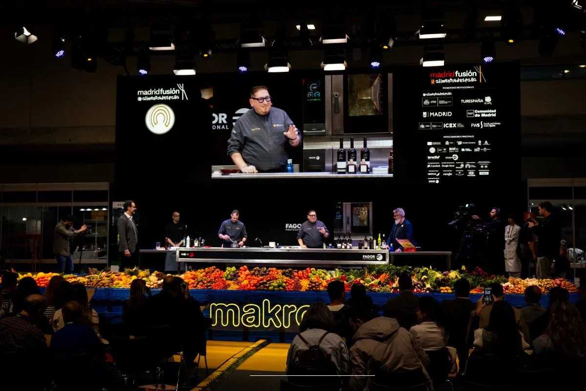 A chef cooking on the Madrid Fusión auditorium stage in front of a seated audience, with crates of vegetables along the front of the stage
