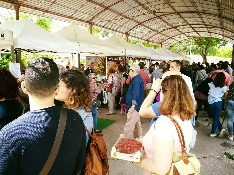 Shoppers browsing white-tented producer stalls under a covered market walkway, one holding a box of strawberries