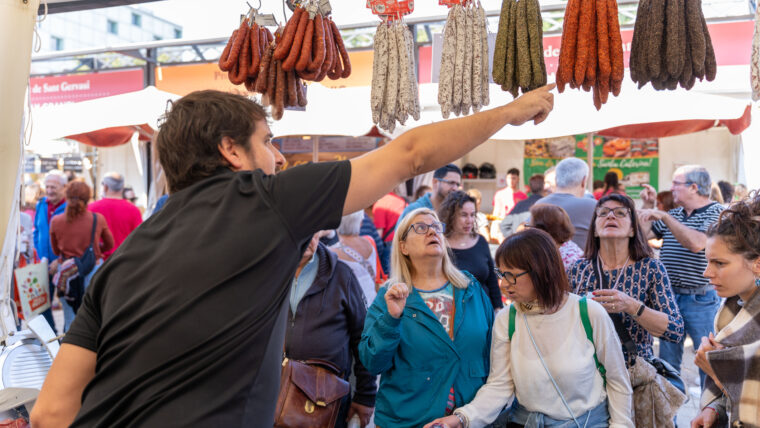 A stallholder points to hanging cured sausages as visitors look on at the 2025 Mercat de Mercats fair.