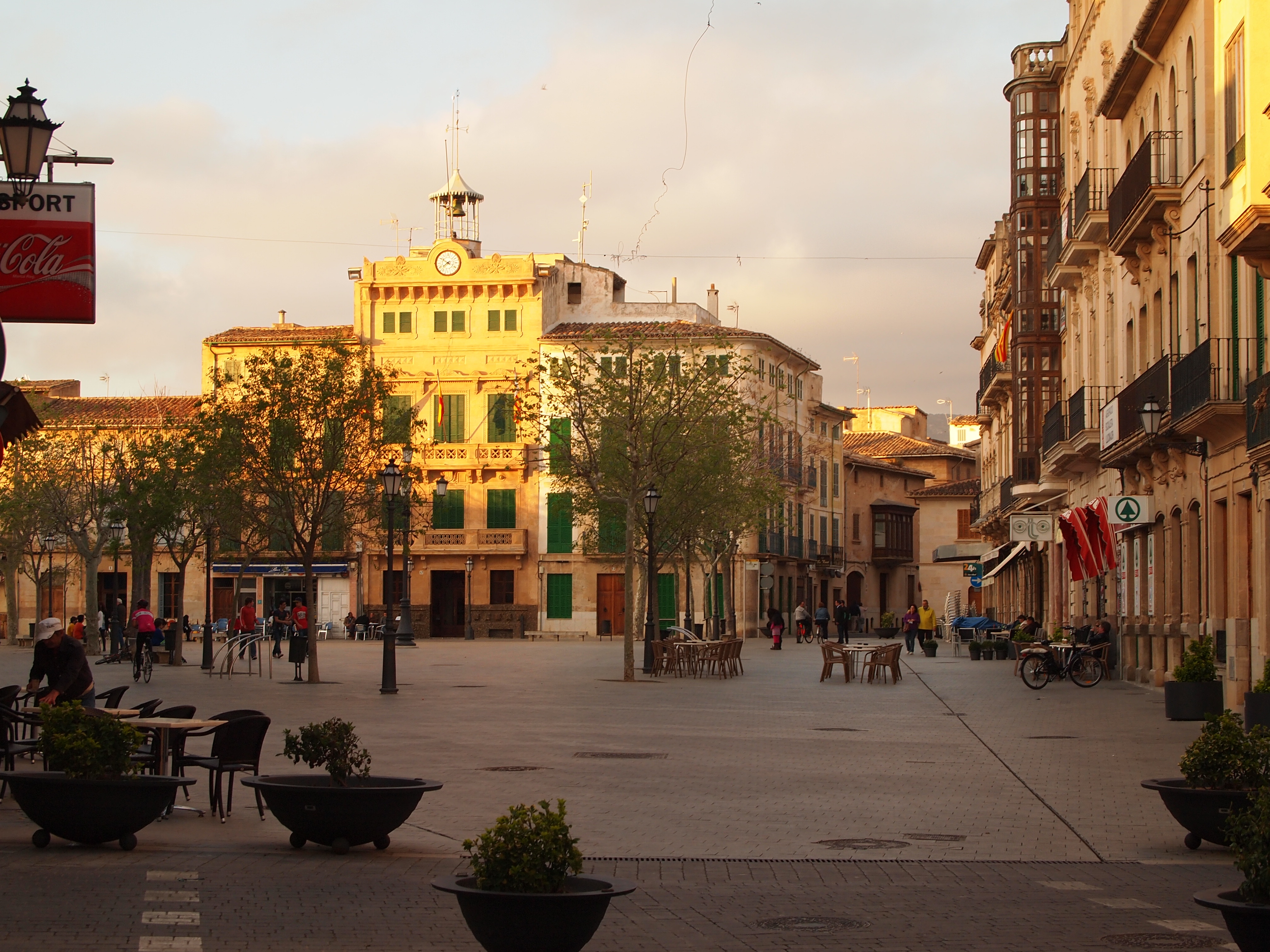 Plaça d'Espanya in Llucmajor at golden hour, with the ornate town hall clock tower, outdoor café tables, cyclists and pedestrians, and a Coca-Cola sign on a shopfront awning
