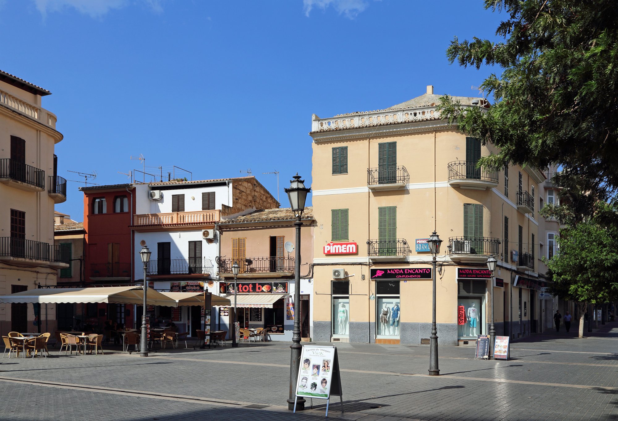 A street corner in Inca's old town at midday, pastel-coloured building façades, a wrought-iron street lamp, and shopfronts including a 'PIMEM' sign, 'Moda Encanto' clothing store and 'tot bo' café under a clear blue sky