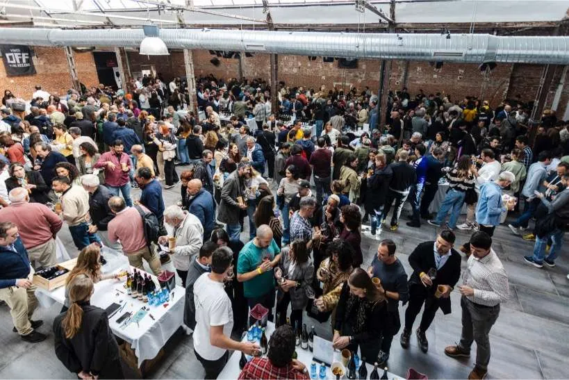 A packed wine fair in a brick-walled industrial hall, visitors tasting at tables of bottles