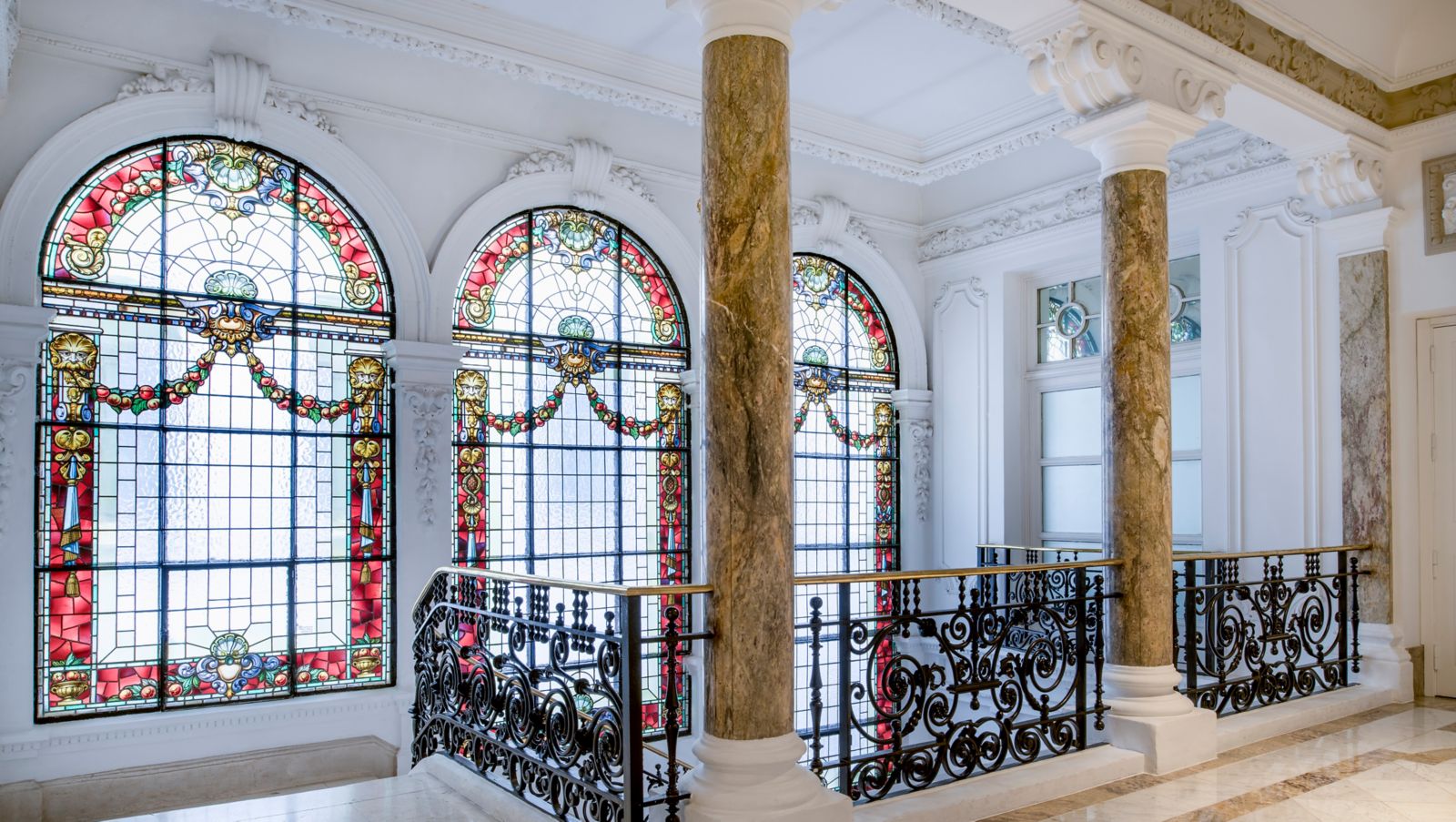A stair landing with three arched stained-glass windows in reds and golds, veined marble columns and a wrought-iron balustrade.
