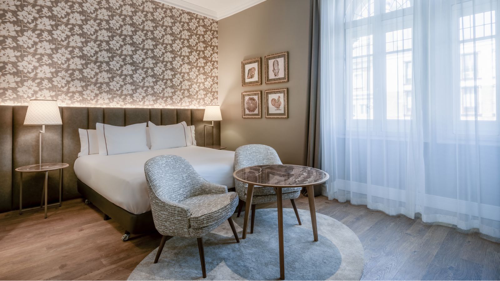 A guest room corner: two patterned armchairs and a round marble-topped table on a circular rug beside the bed, with sheer curtains over tall windows.