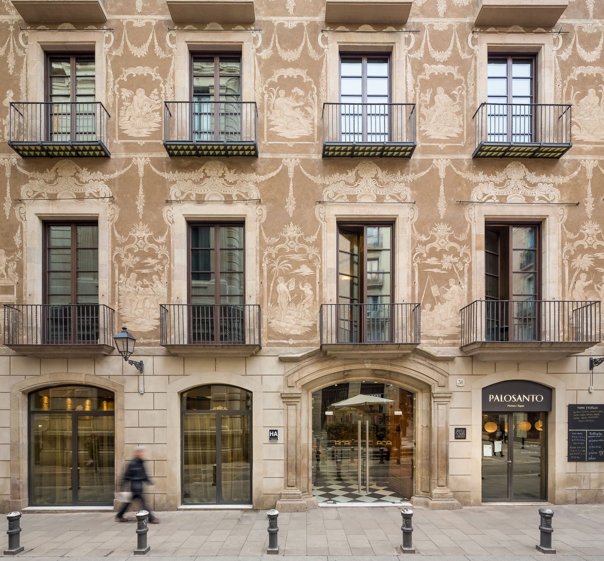 Ornate stucco facade of Arai Aparthotel on a Barcelona street, with wrought-iron balconies and the Palosanto restaurant signage at street level