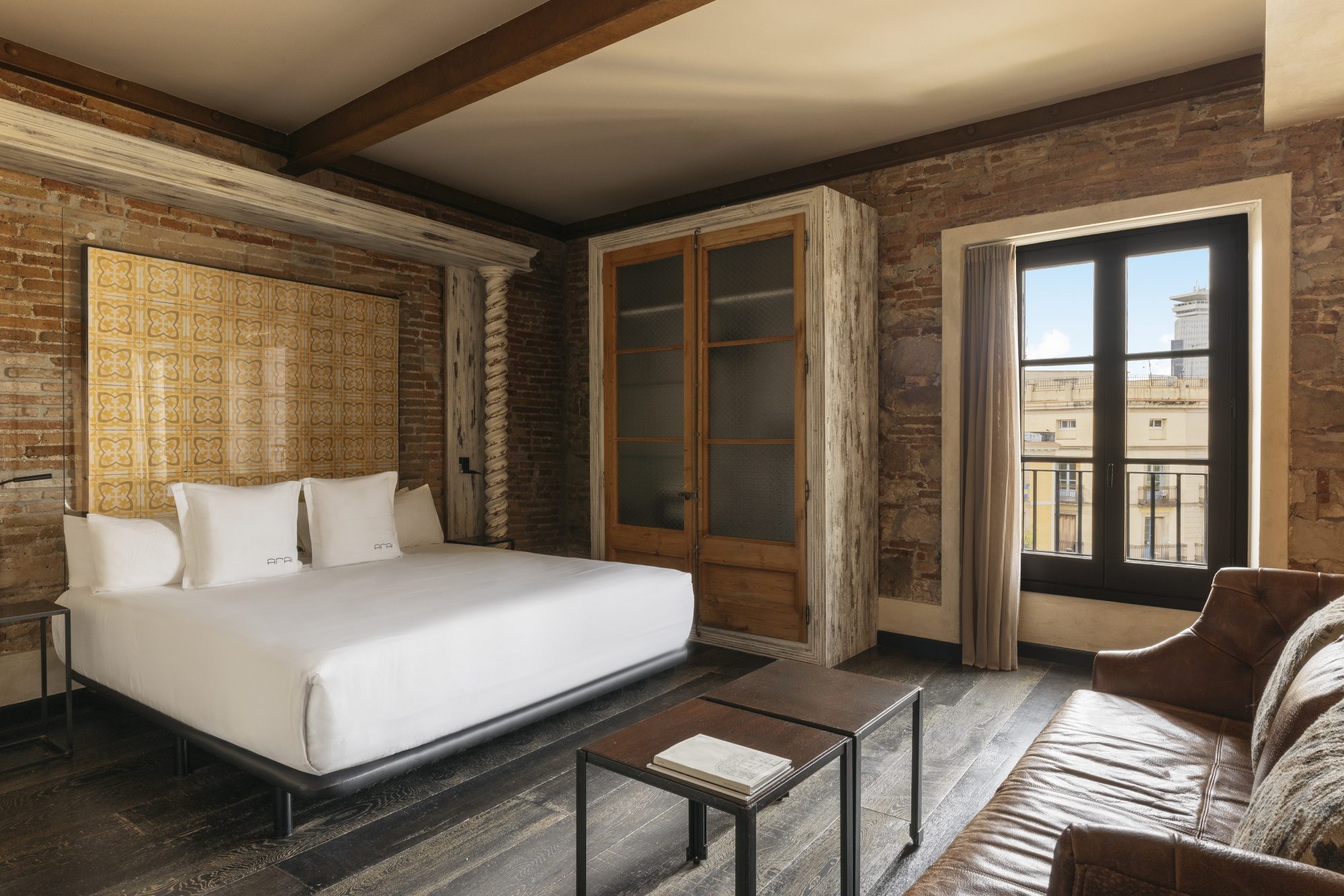 Guest room at Arai Aparthotel with exposed brick walls, dark wood ceiling beams, a white bed against a patterned headboard, and a leather armchair by the balcony window