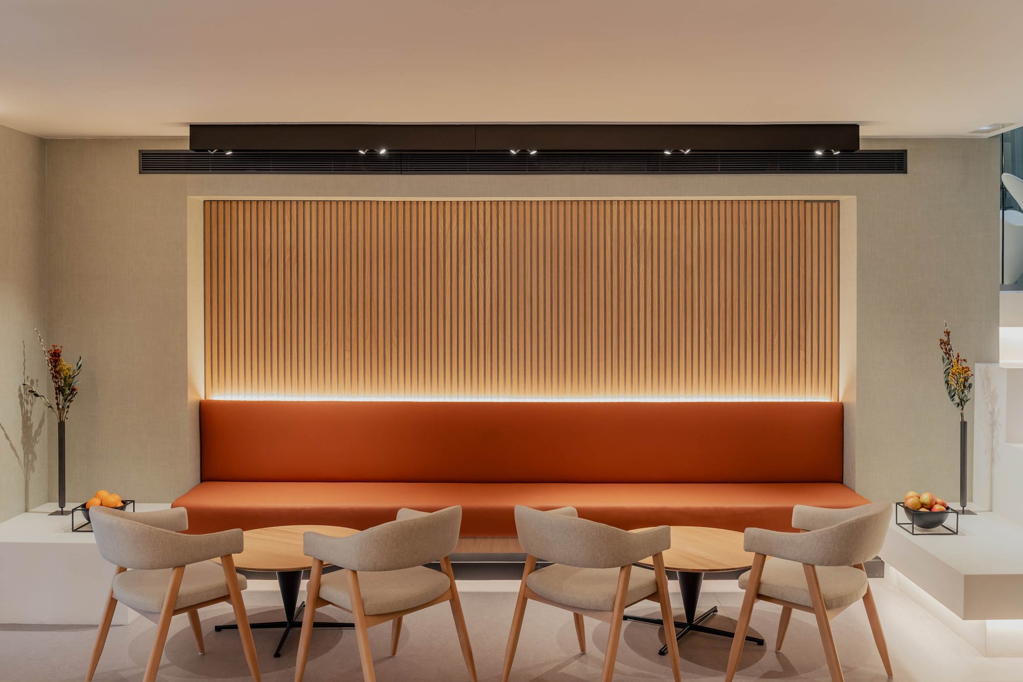 Lounge at Hotel Arconte with a long rust-coloured banquette, round tables, pale chairs and a slatted wood panel lit from above