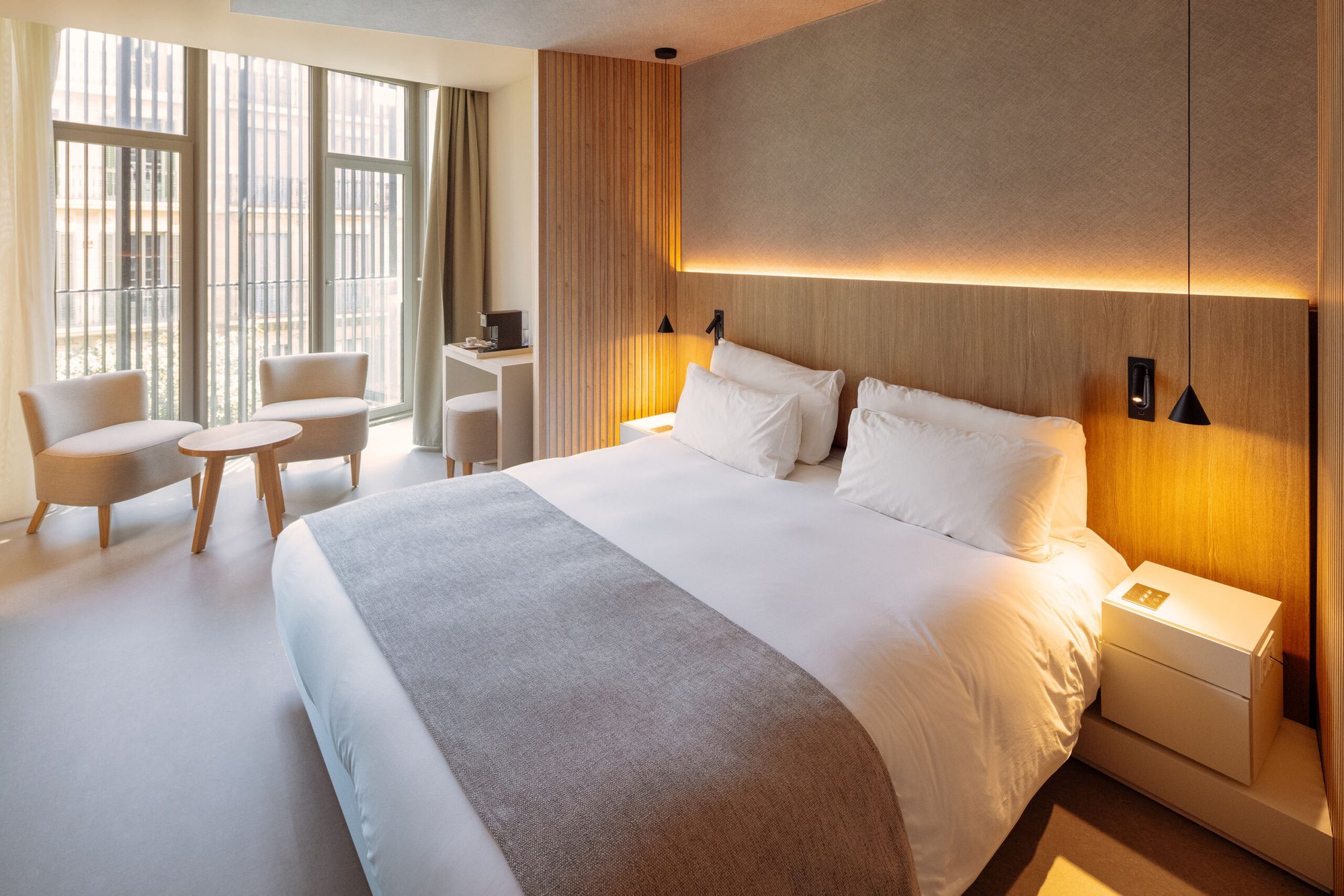 Guest room at Hotel Arconte with a wood-panelled headboard wall, white bedding with a grey throw and two armchairs by tall windows