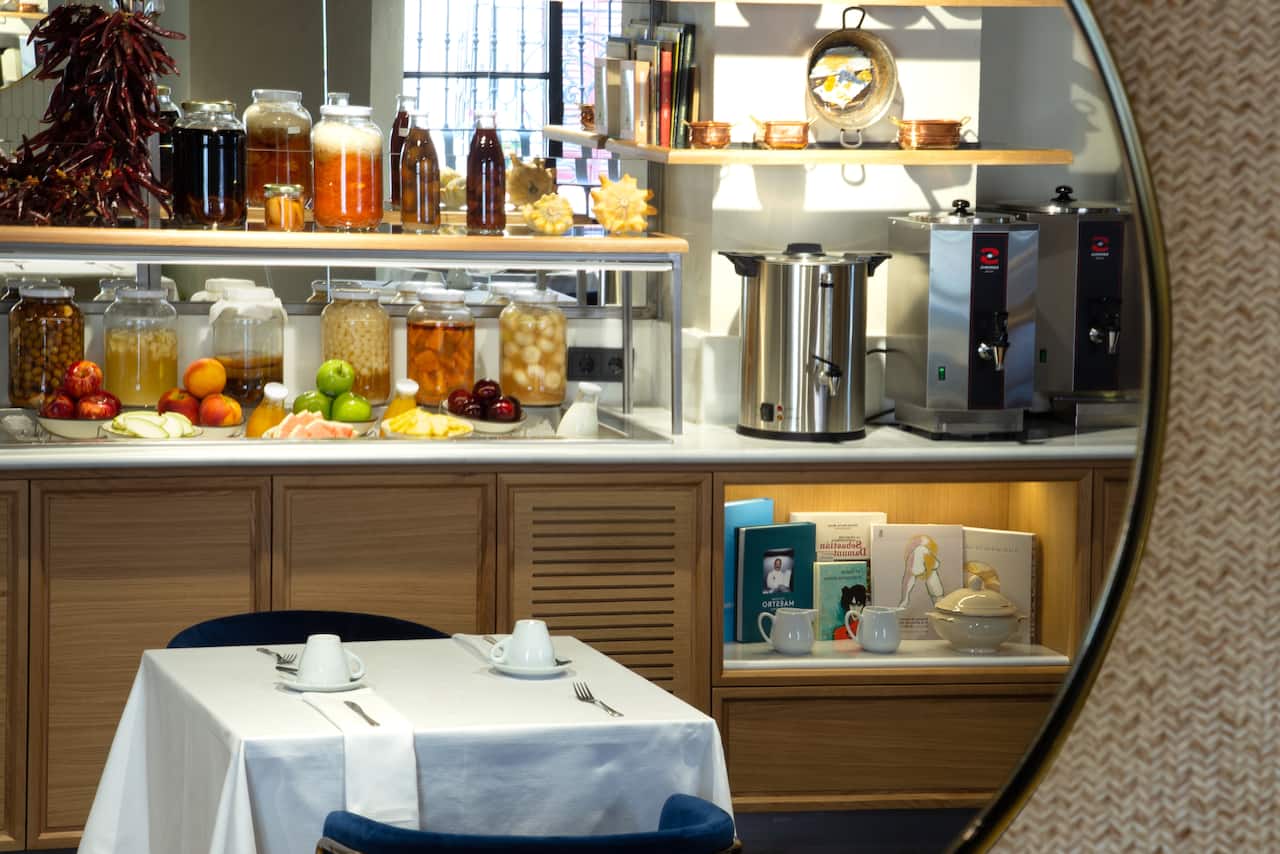 A breakfast buffet counter with jars of preserves, fresh fruit and drink dispensers, reflected in a round mirror, with a table set for two.