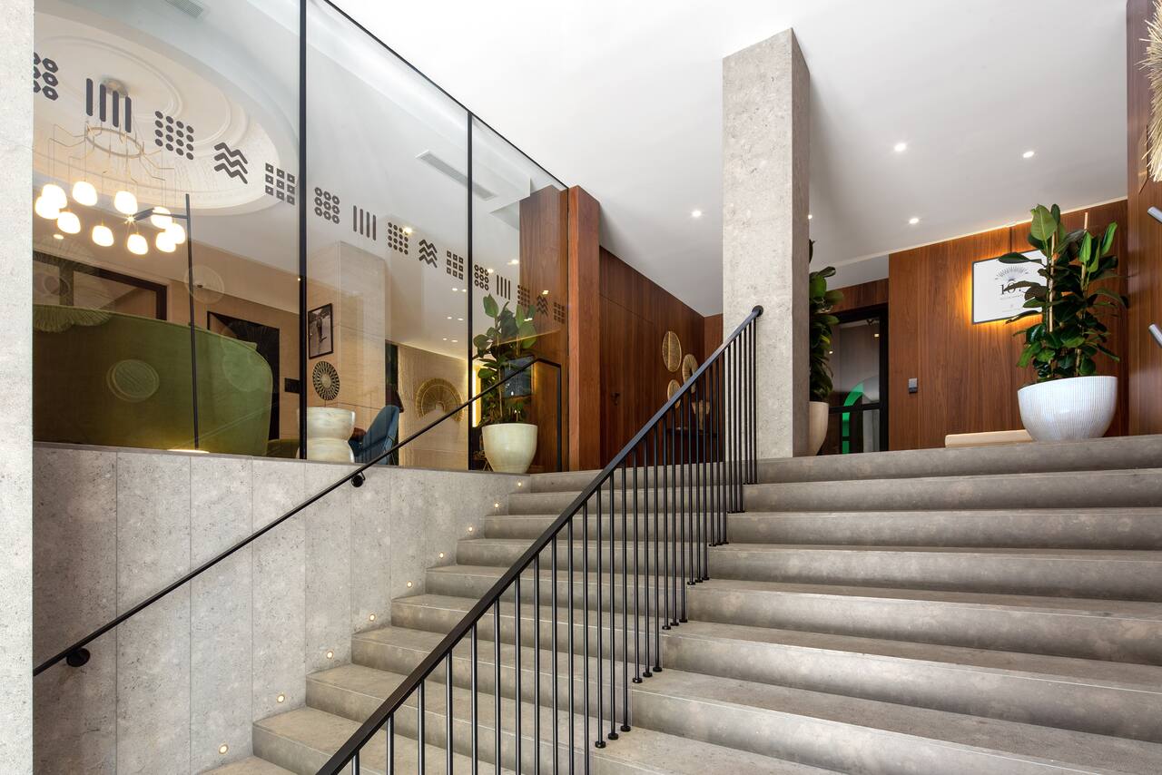 A hotel staircase in stone and dark wood, leading up to a lobby seating area visible through glass panels.