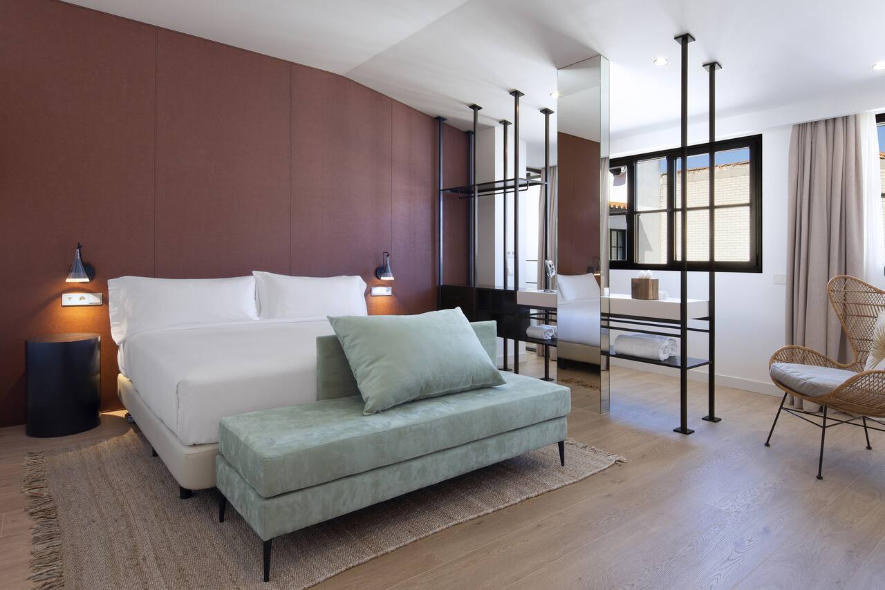 A hotel room with a terracotta headboard wall, a green velvet bench at the foot of a white bed, a wicker chair and an open glass-walled bathroom area.