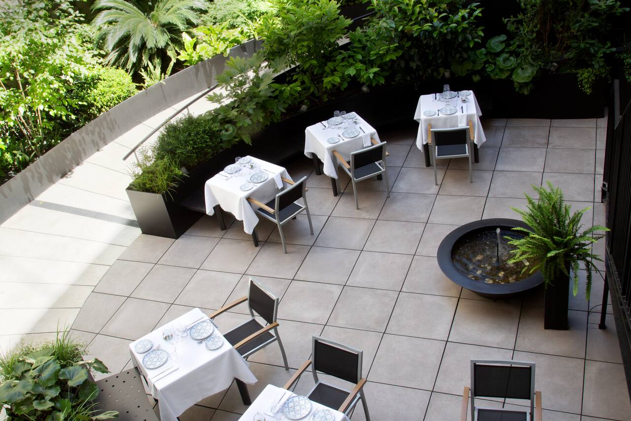 An outdoor restaurant terrace with white-clothed tables set for dining, surrounded by dense greenery and a small water feature.