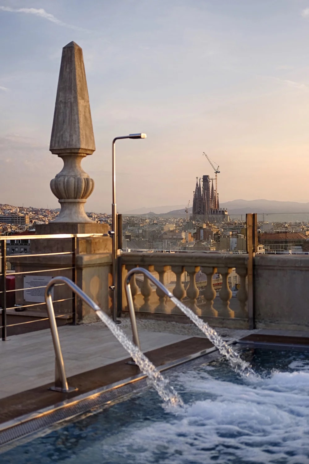The hotel's rooftop pool with steel water jets and a stone balustrade, the Sagrada Familia visible in the distance.