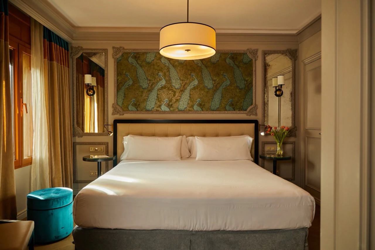 A hotel bedroom with a king bed, a teal velvet ottoman, and a green peacock-patterned panel above the headboard.