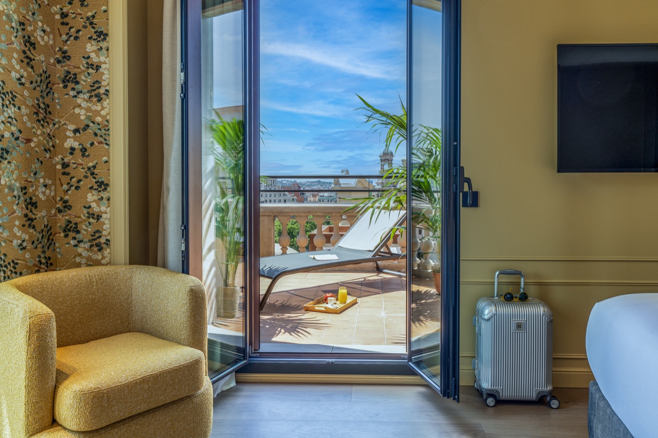 A guest room with glass doors open onto a private terrace furnished with a lounge chair and a breakfast tray.
