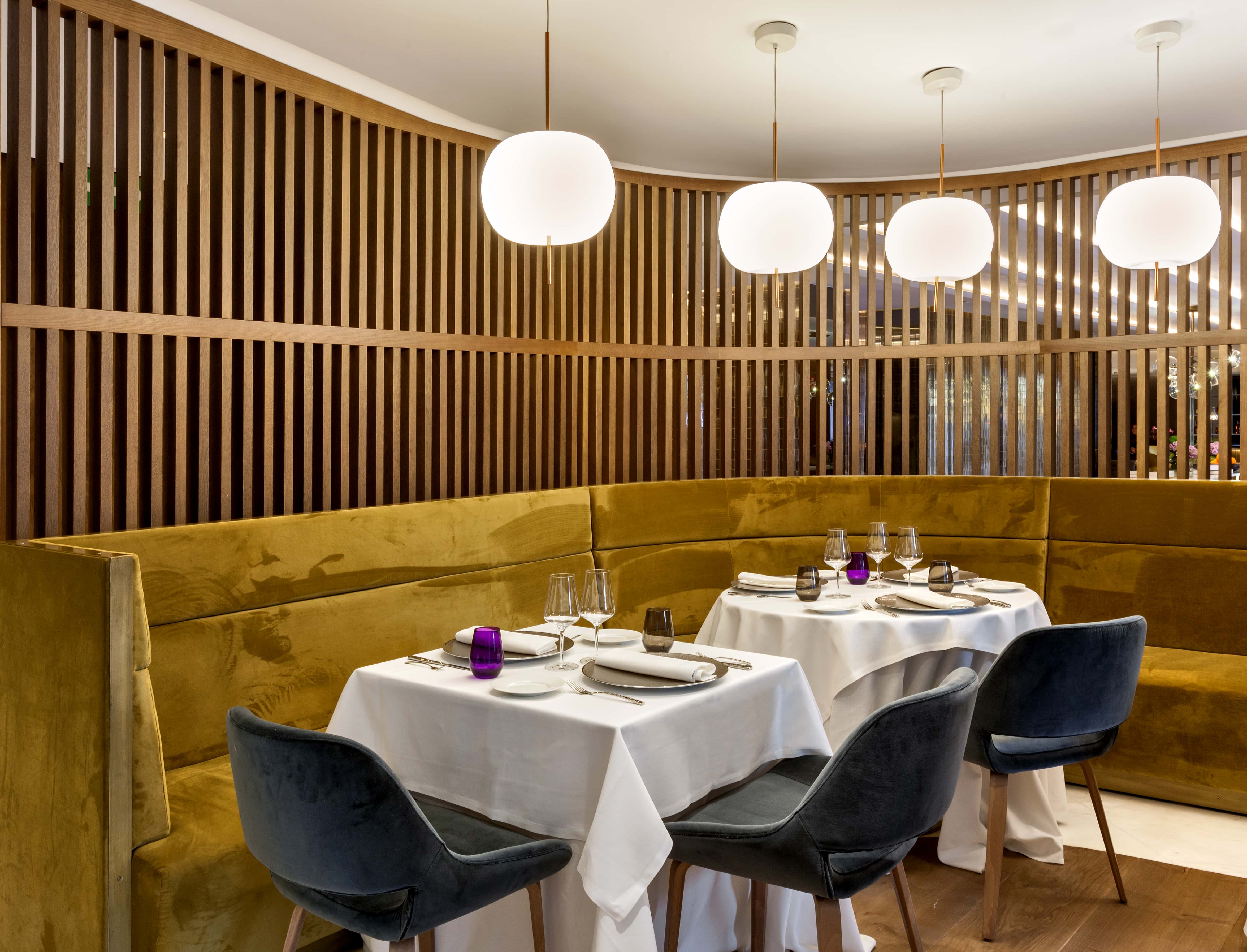 The dining room at Barcelo Emperatriz, with two tables laid in white linen and set with wine glasses along a mustard velvet banquette, beneath a slatted oak screen and white globe pendants.