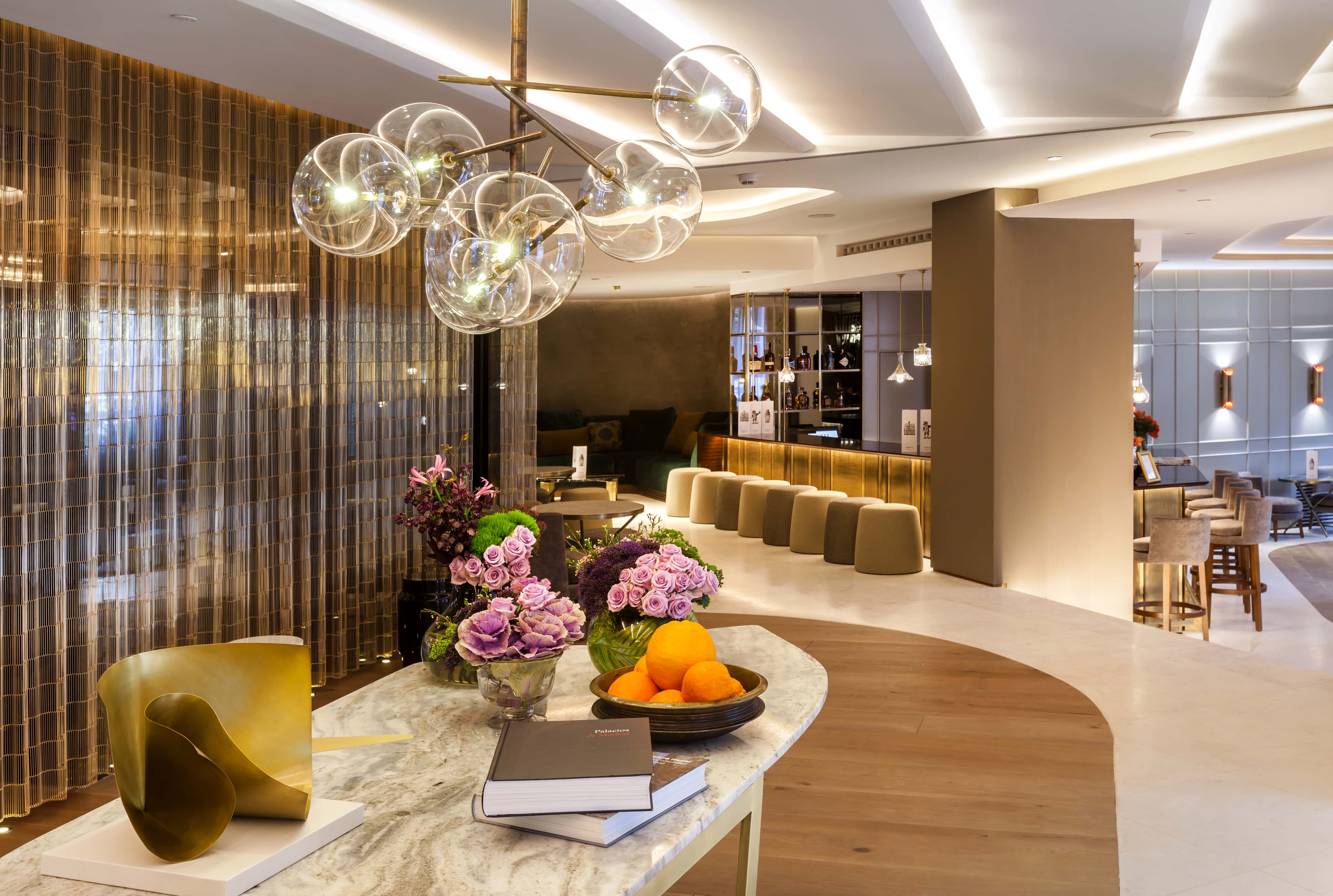 The lobby lounge at Barcelo Emperatriz: a marble console holding flower arrangements, a bowl of oranges and a brass sculpture, with a glass-globe chandelier overhead and the bar counter and stools beyond.