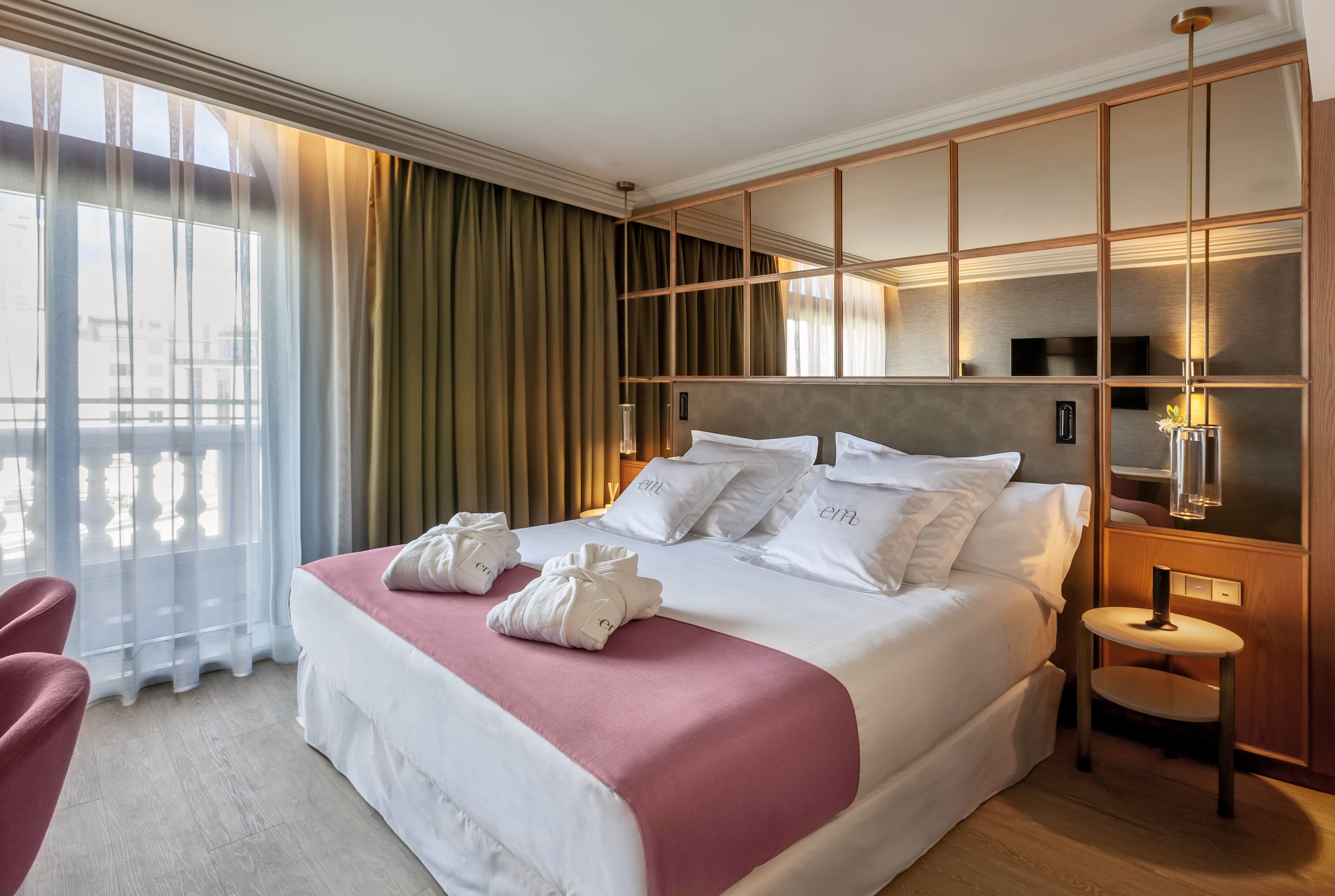 A balconied guest room at Barcelo Emperatriz with a pink throw and two folded robes across the bed, 'em' monogrammed cushions, and full-height windows onto a stone balustrade.