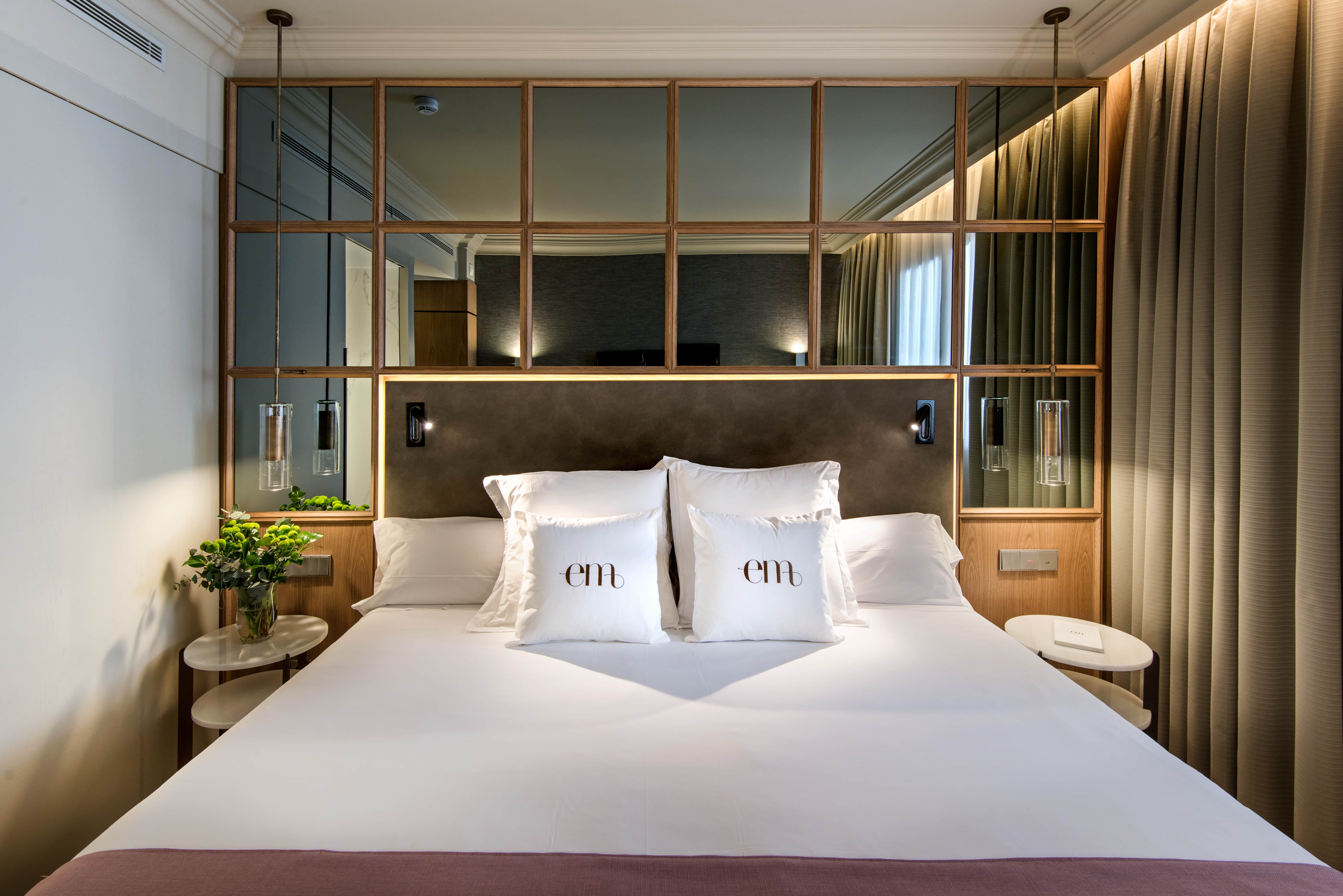 A guest room at Barcelo Emperatriz, the bed made up in white linen with cushions embroidered 'em' beneath an oak-framed mirrored panel and hanging glass reading lights.