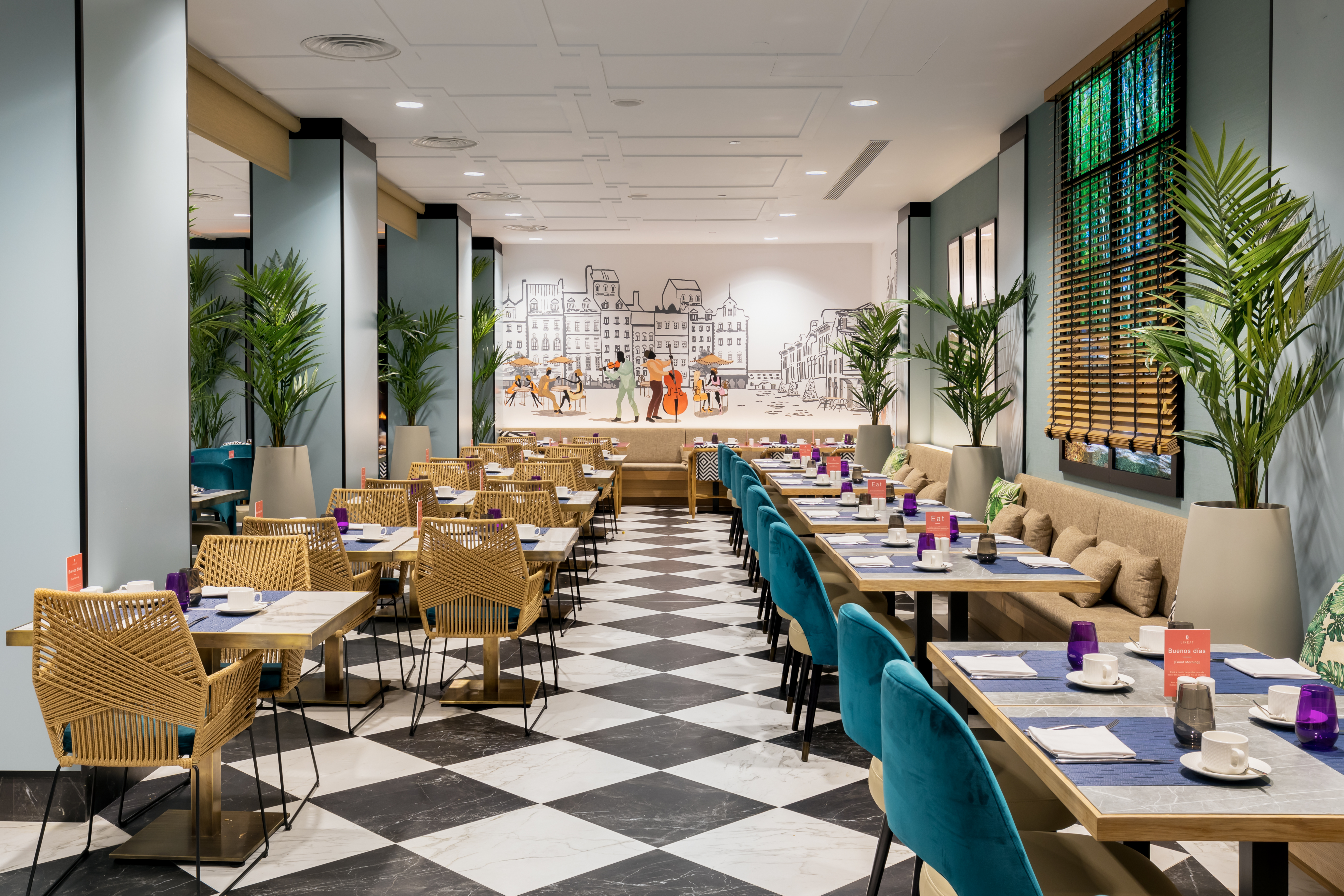 The restaurant at Barcelo Imagine: rattan and teal chairs at tables laid with cups and napkins on a black-and-white chequerboard floor, beneath a line-drawn townscape mural and tall potted palms.