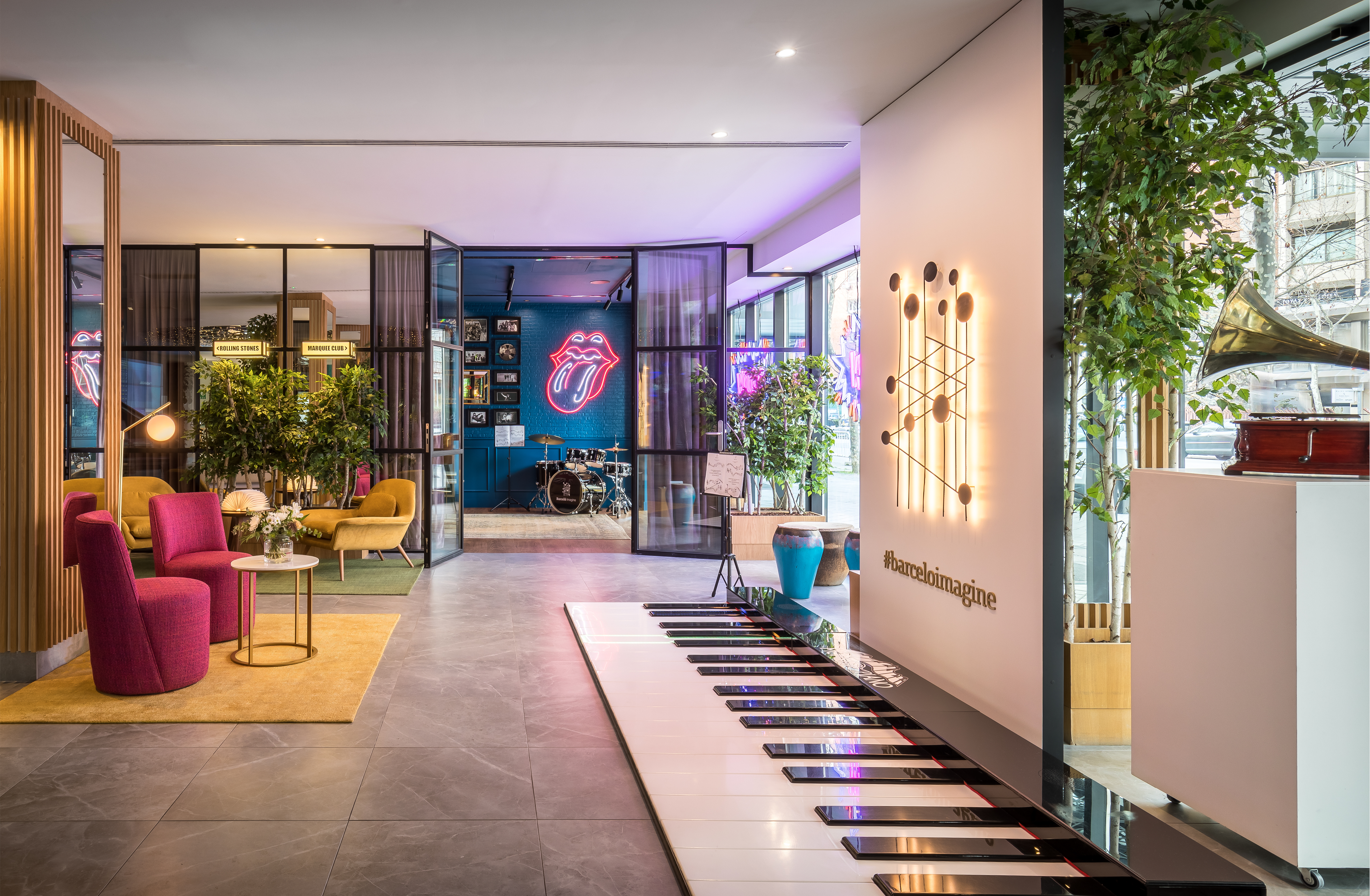 The lobby at Barcelo Imagine, with an oversized piano-keyboard installation set into the floor, pink and mustard armchairs on rugs, and a glass-walled room beyond holding a drum kit under a neon lips sign.