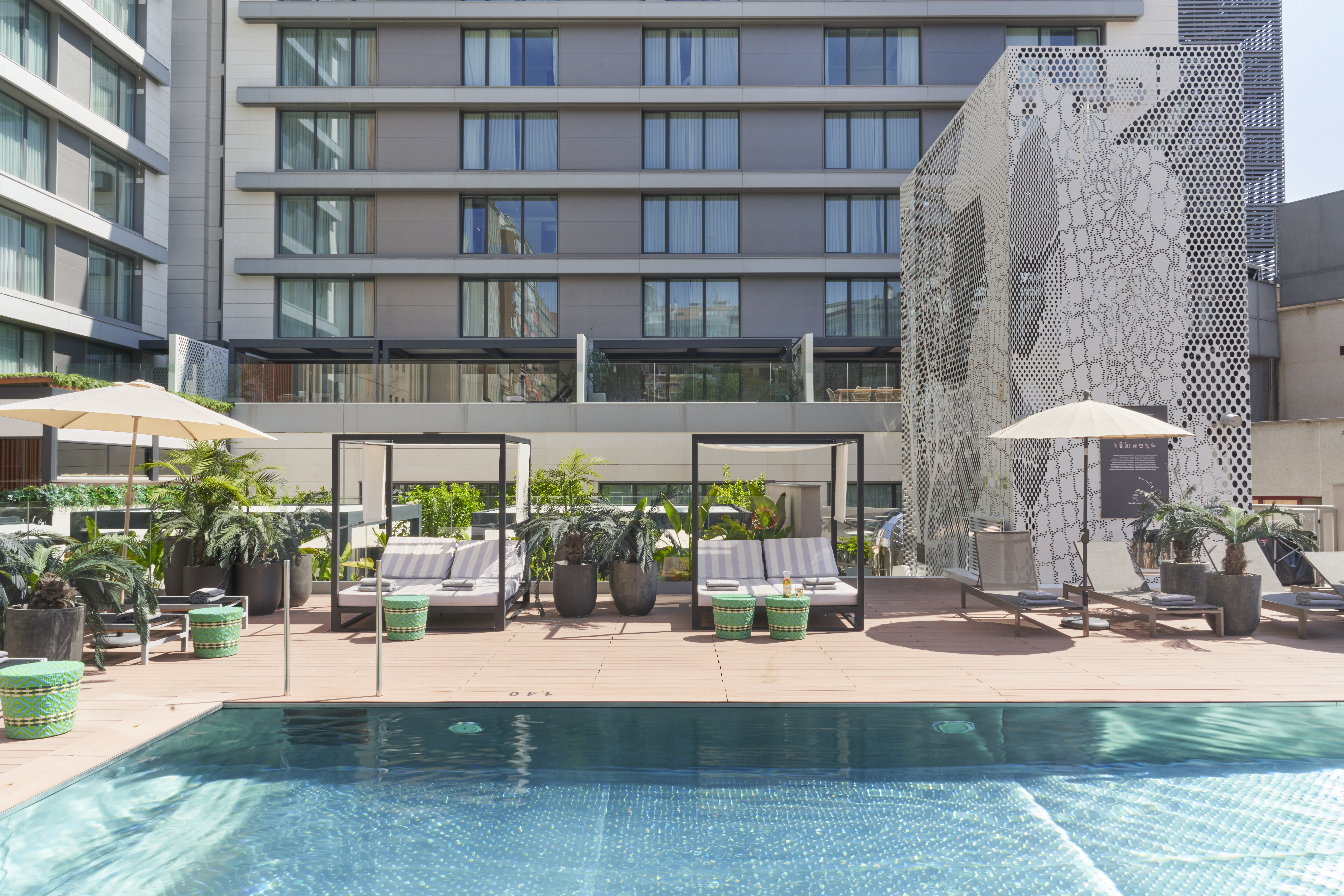 The outdoor pool at Barcelo Imagine, with parasols, striped loungers under canopied beds and green woven side tables on the decking, and the hotel's balconied facade rising behind.