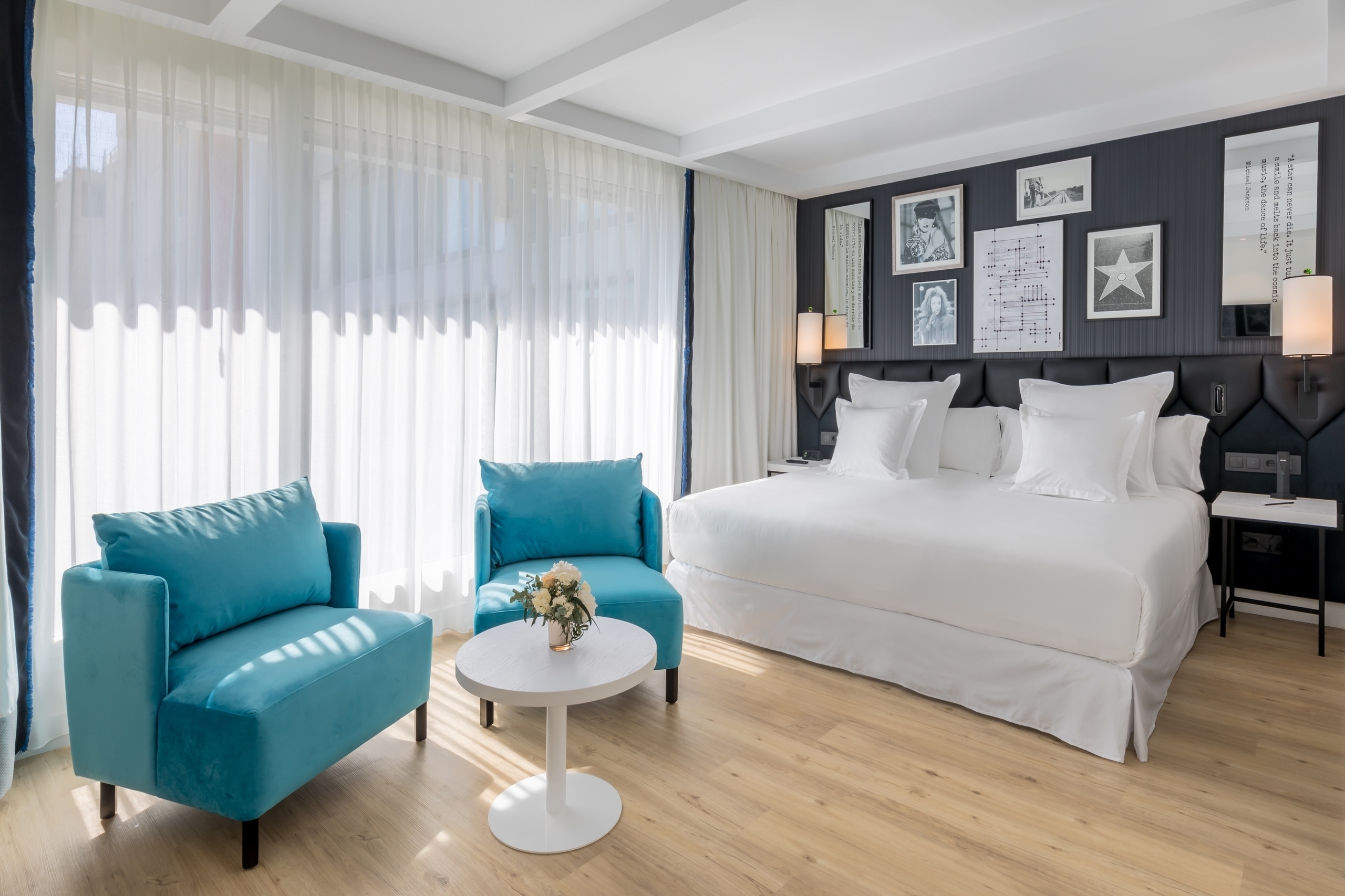 A guest room at Barcelo Imagine with a white-linen bed against a dark padded headboard wall hung with framed black-and-white music prints, and two teal armchairs by the full-height curtained window.