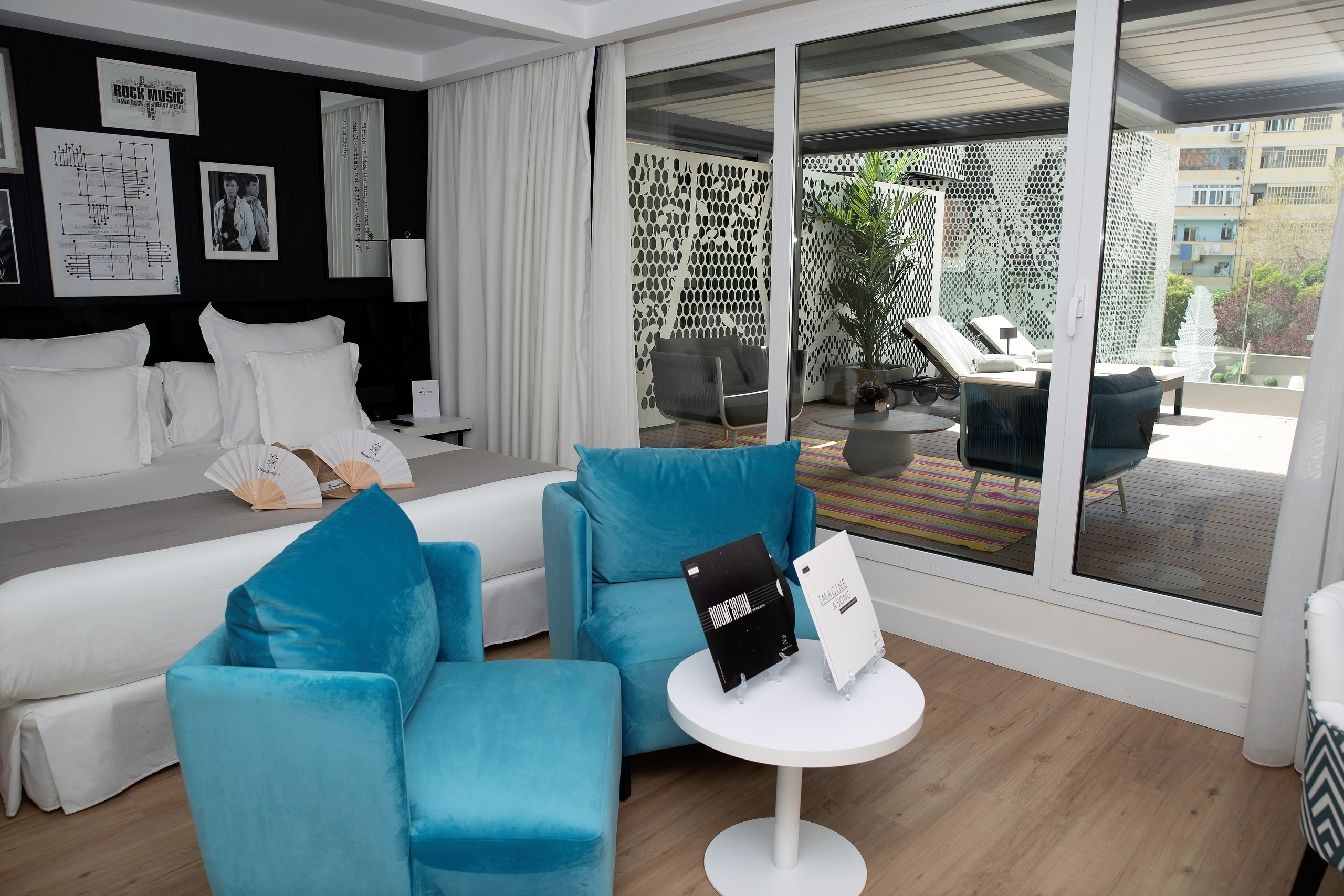 A suite at Barcelo Imagine, its bed and teal armchairs beside sliding glass doors opening onto a private terrace with loungers, screened by white perforated panels.