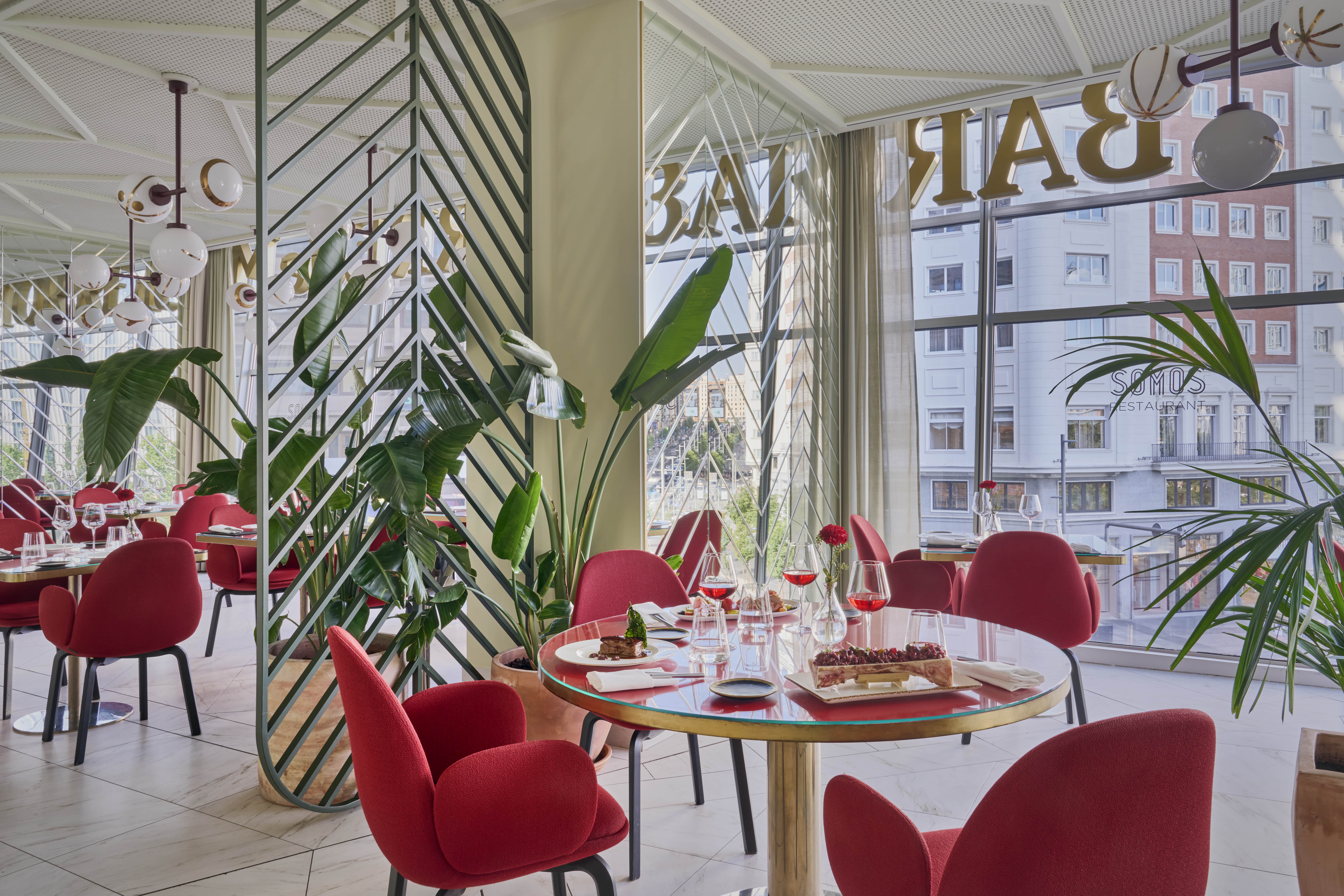 The restaurant at Barcelo Torre de Madrid: round tables with red upholstered armchairs and laid plates beside floor-to-ceiling windows carrying gold 'BAR' lettering, with large-leafed plants between the tables.