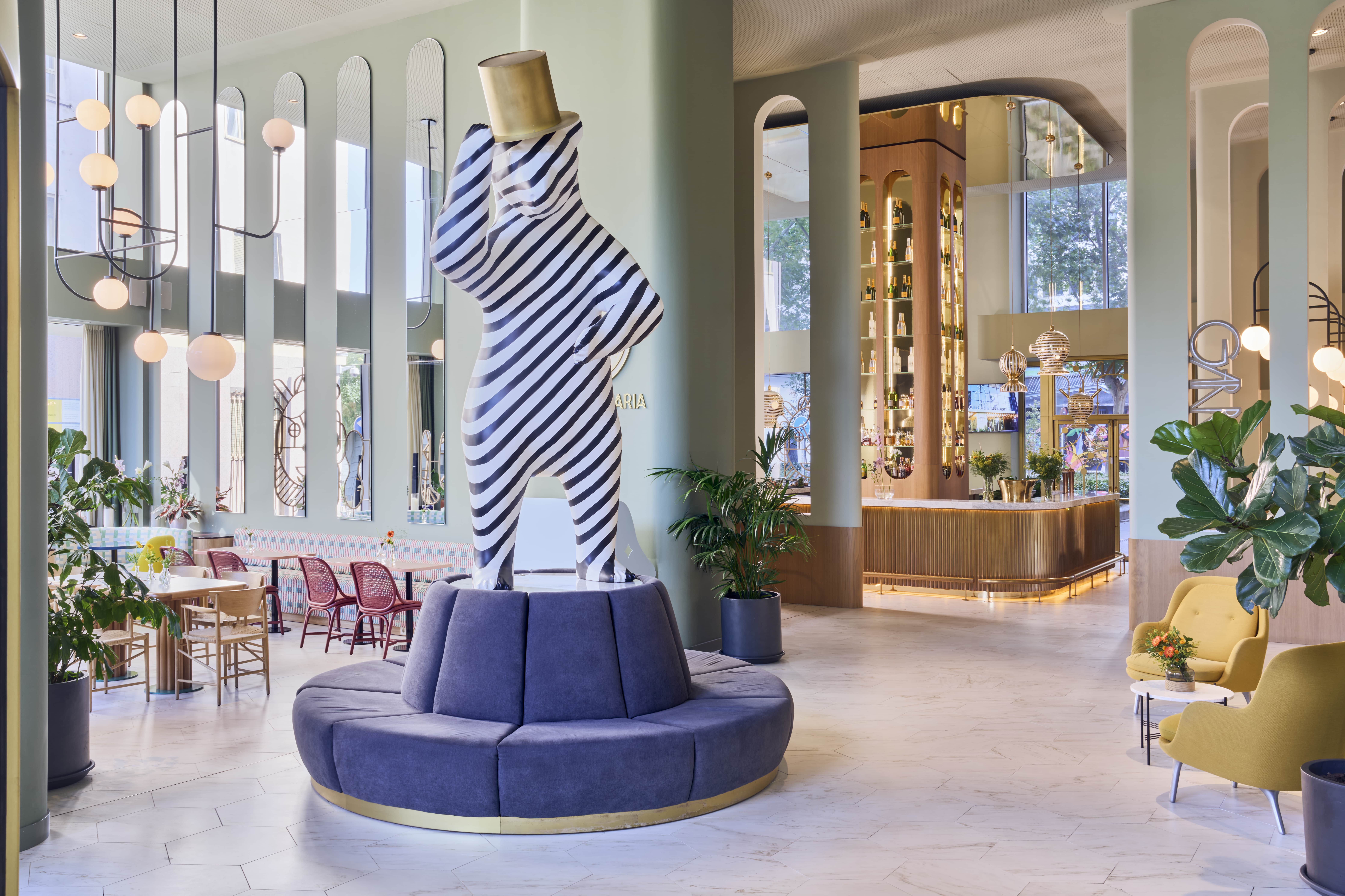 The lobby at Barcelo Torre de Madrid, where a black-and-white striped figure in a brass top hat stands on a circular blue banquette between mint-green columns, with the brass-fronted bar counter beyond.