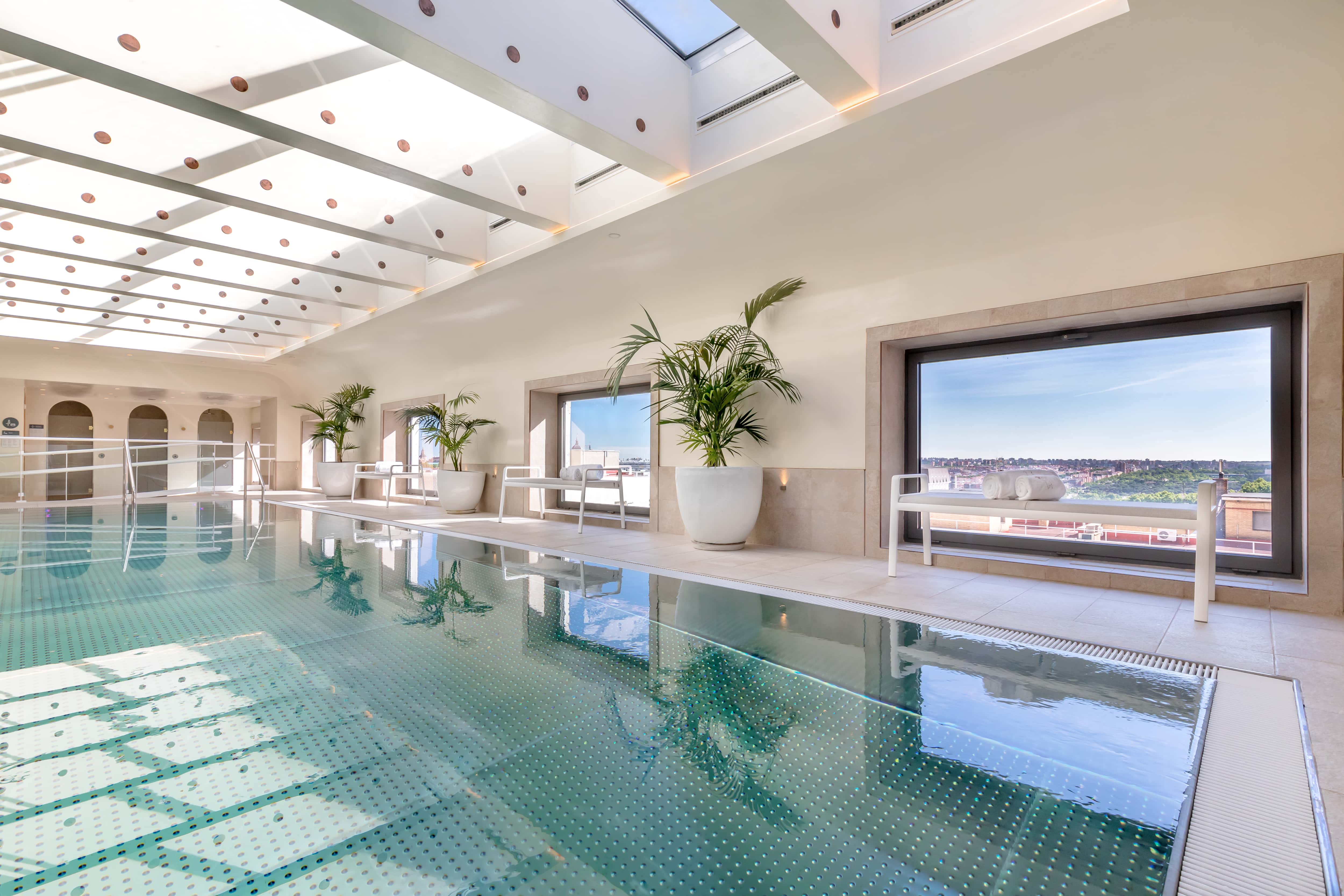 The indoor pool at Barcelo Torre de Madrid, a long tiled pool under a slatted skylight, with potted palms and loungers along a picture window looking out over rooftops and greenery.