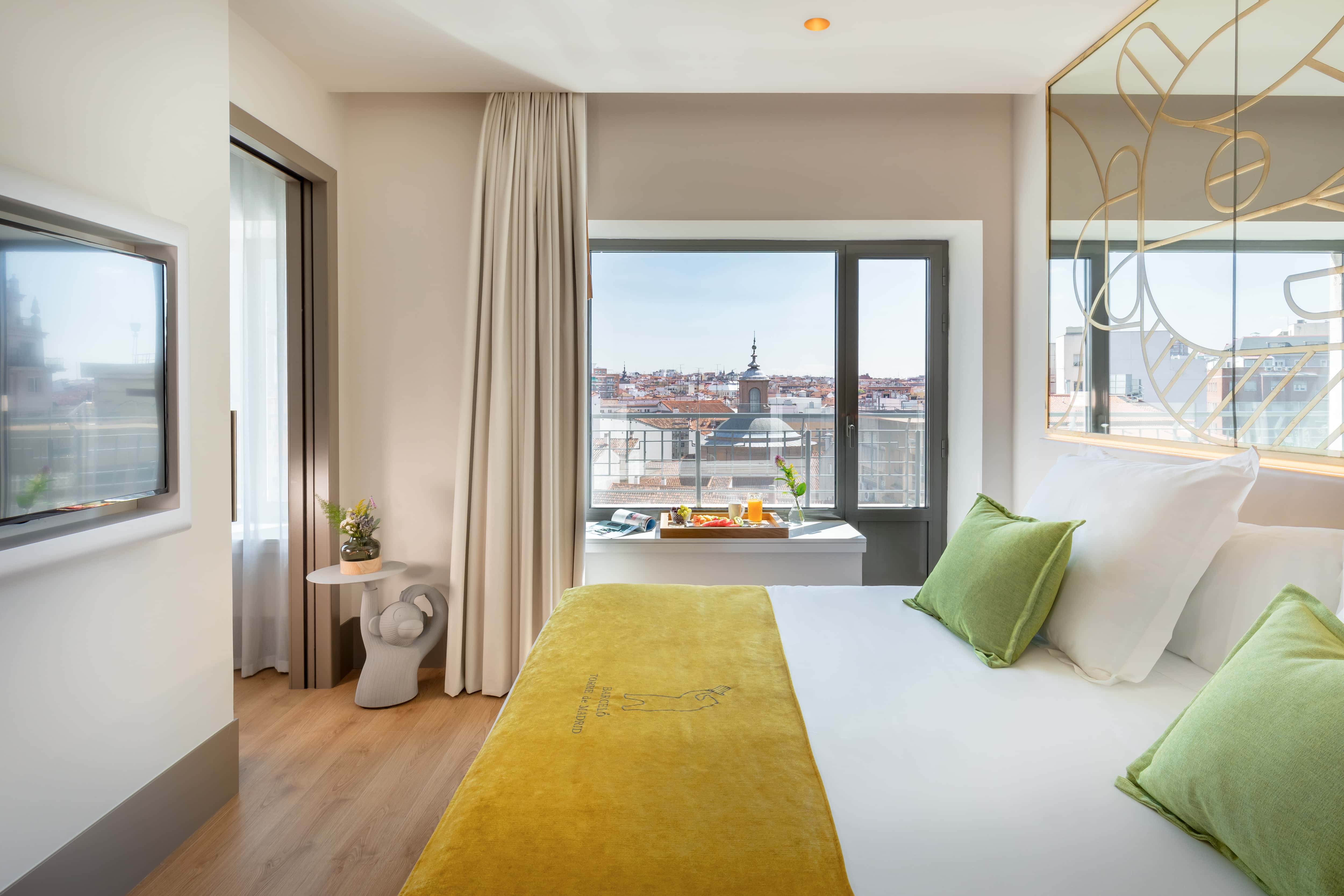 A guest room at Barcelo Torre de Madrid with a yellow embroidered throw across the bed, green cushions, a gold line-drawn mirrored headboard panel and French doors onto a balcony over Madrid rooftops.