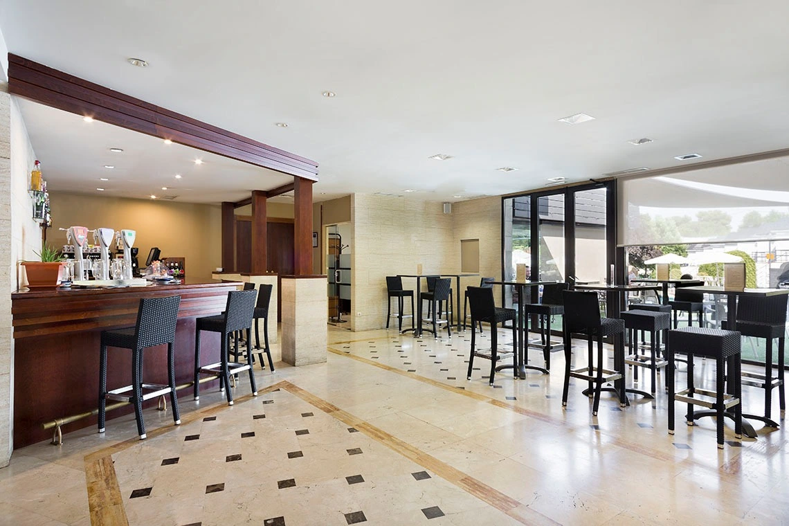 Hotel bar with a dark wood counter and beer taps, black bar stools, and a row of high-top tables leading toward glass doors to the terrace.