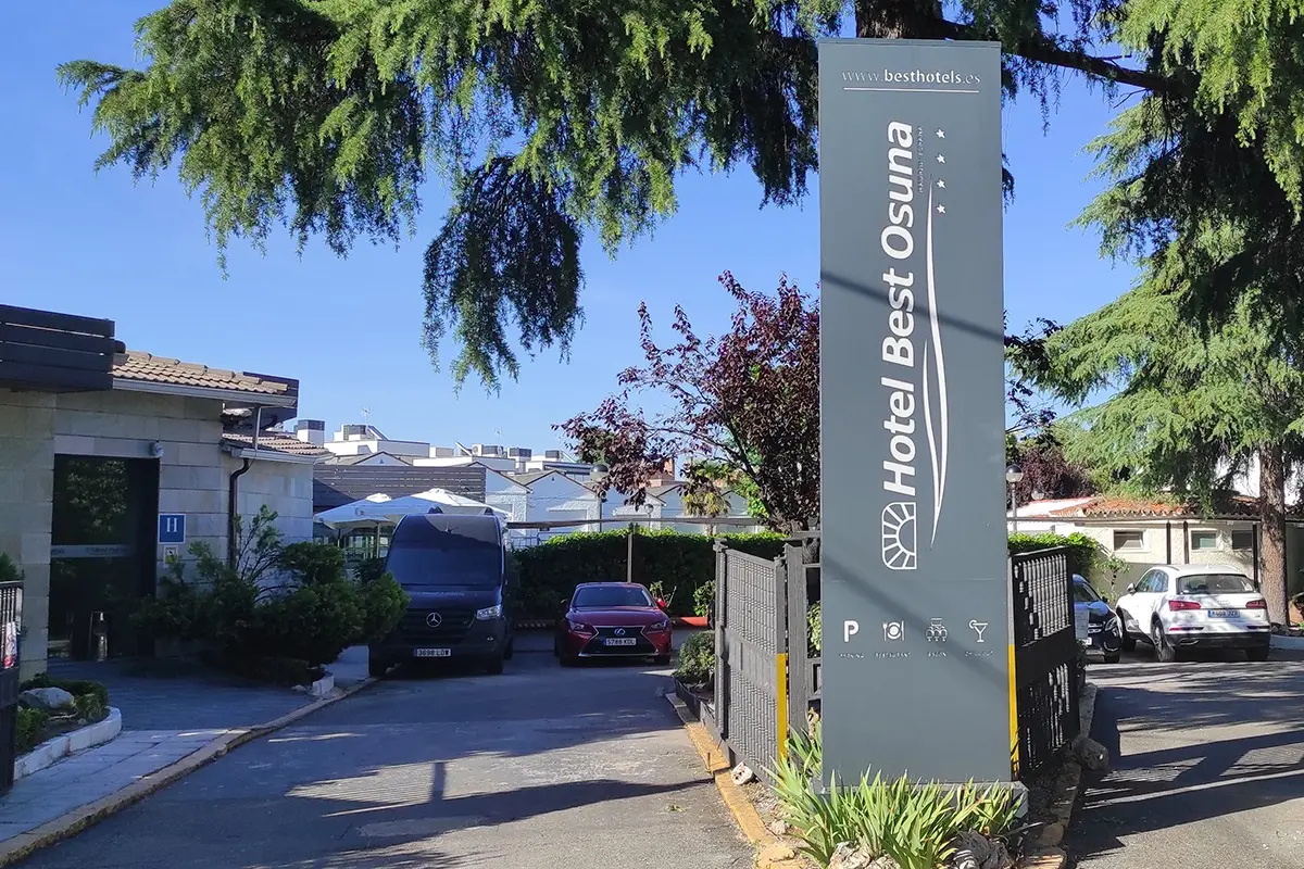 Grey monolith sign reading Hotel Best Osuna with its four-star mark, at the hotel's driveway entrance with parked cars and trees.