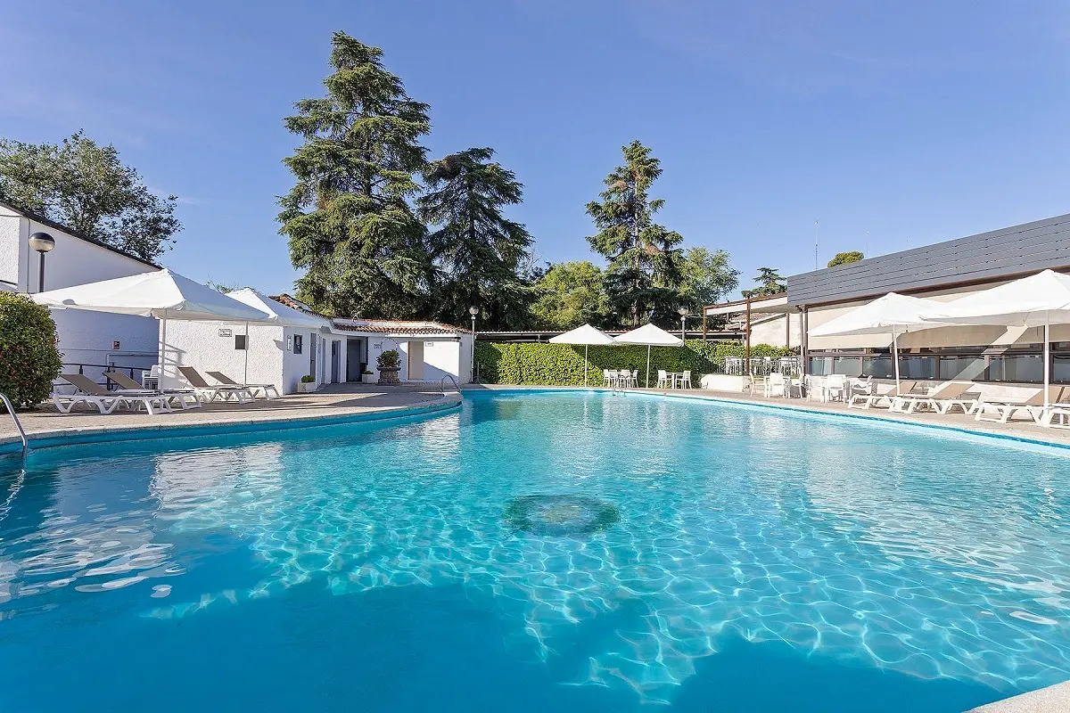 Large outdoor swimming pool surrounded by white sun loungers and umbrellas, low white poolside buildings, and tall pine trees behind.
