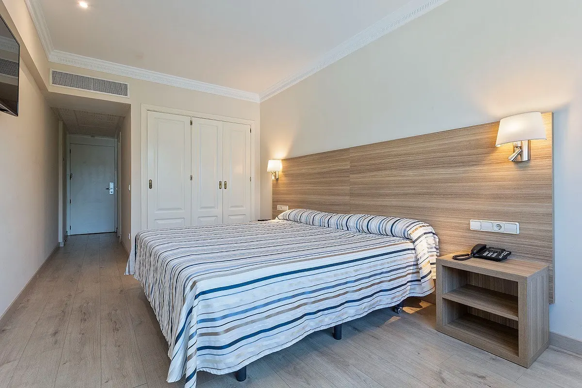 Hotel room with a striped blue and white bedspread, a light wood headboard wall, a bedside shelf unit with a telephone, and a built-in wardrobe.