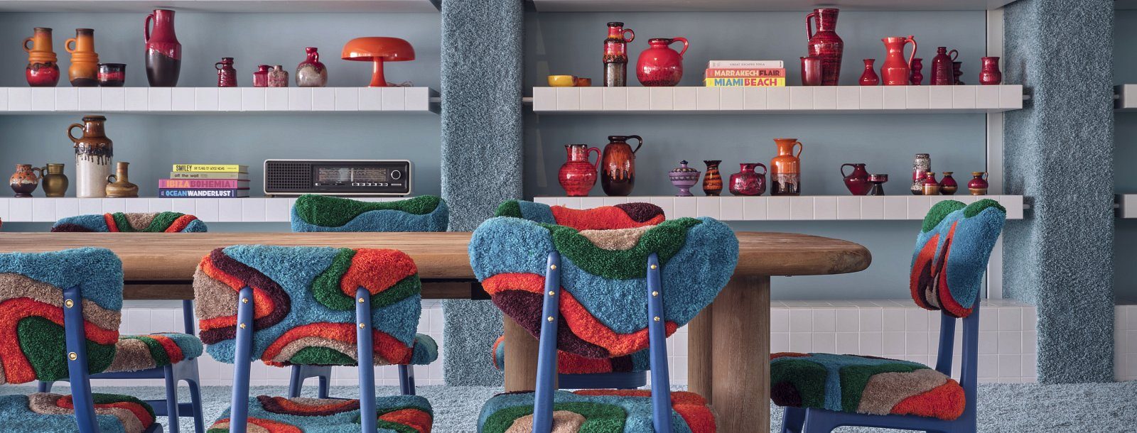Retro-styled lounge at Bikini Island & Mountain Hotel Es Trenc with patterned tufted chairs around a wood table and shelves of colorful ceramic vases