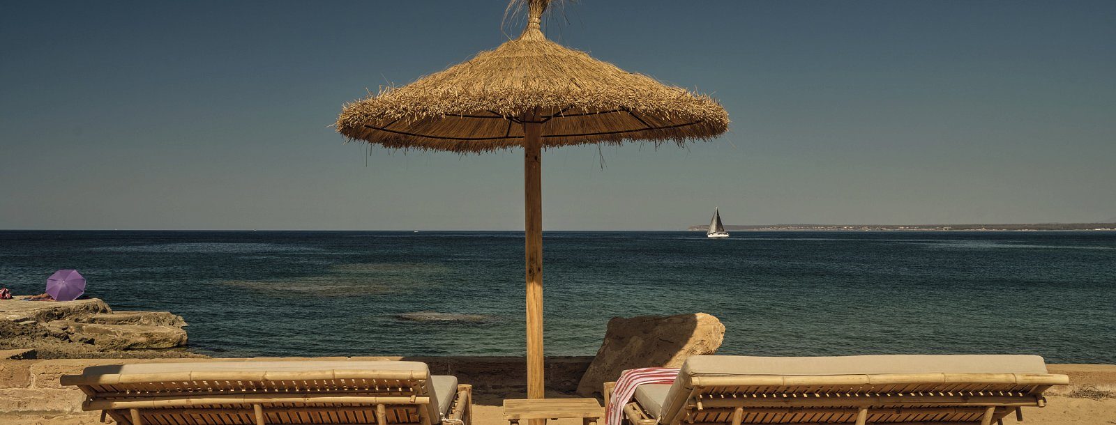 Beachfront sun loungers under a straw parasol facing the sea at Bikini Island & Mountain Hotel Es Trenc, with a sailboat on the horizon