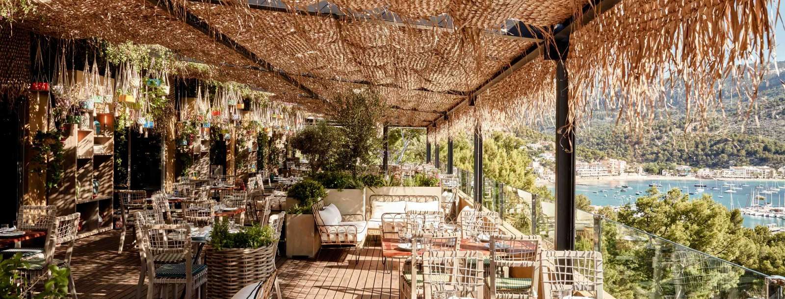 Neni restaurant terrace at Bikini Island & Mountain under a woven rattan pergola hung with plants, wicker chairs set for dining with a view over Port de Sóller harbour